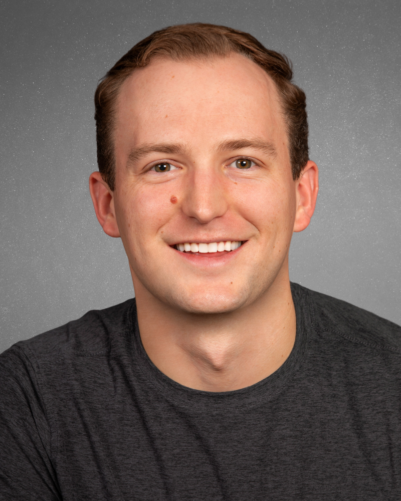 A smiling young man with short brown hair, light skin, wearing a dark gray shirt against a gray, starry background.