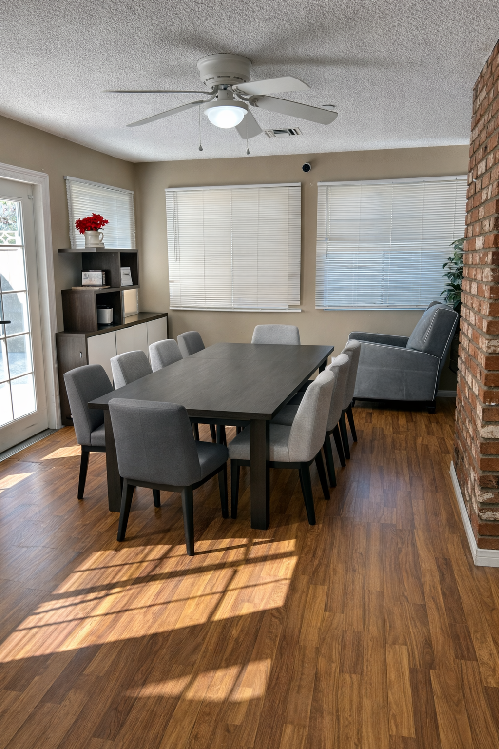Dining room with a long black table surrounded by gray chairs, a ceiling fan, a brick wall, a wooden cabinet with a plant and decorative items, three windows with blinds, a glass door, a gray armchair, and hardwood floors.