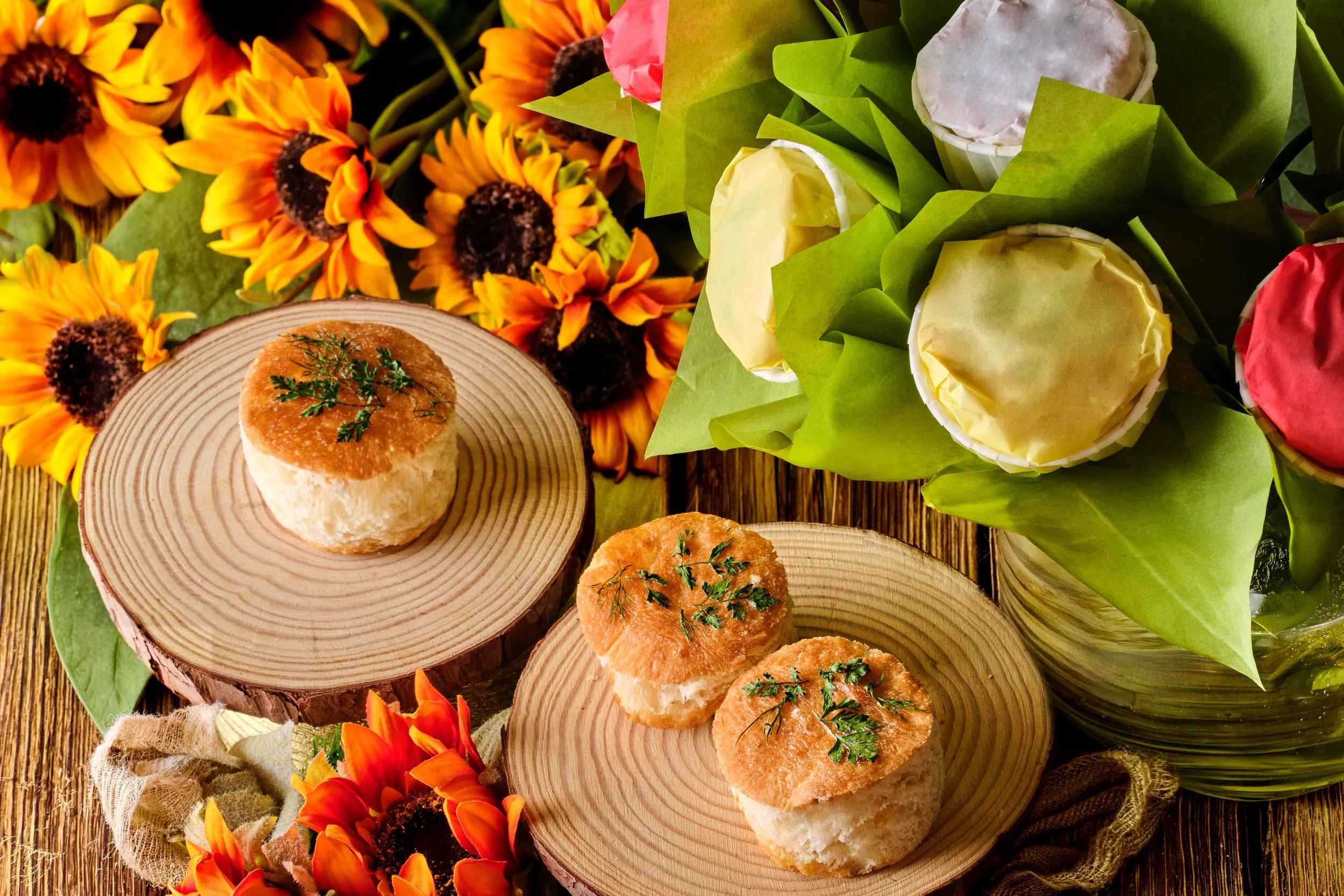 Cupcakes topped with herbs on wooden slabs surrounded by sunflowers and a bouquet of flowers on a wooden table.