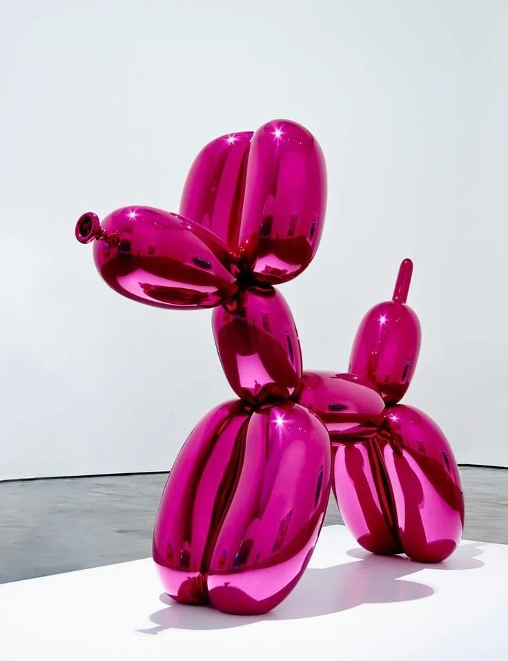 A shiny, pink balloon sculpture of a dog, standing on a white surface in a gallery setting.