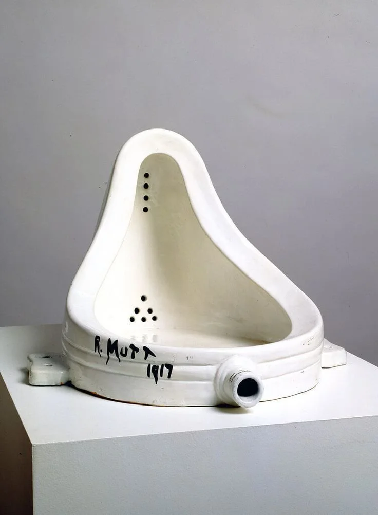 White vintage urinal with black signature and date, mounted on a white pedestal against a gray wall.