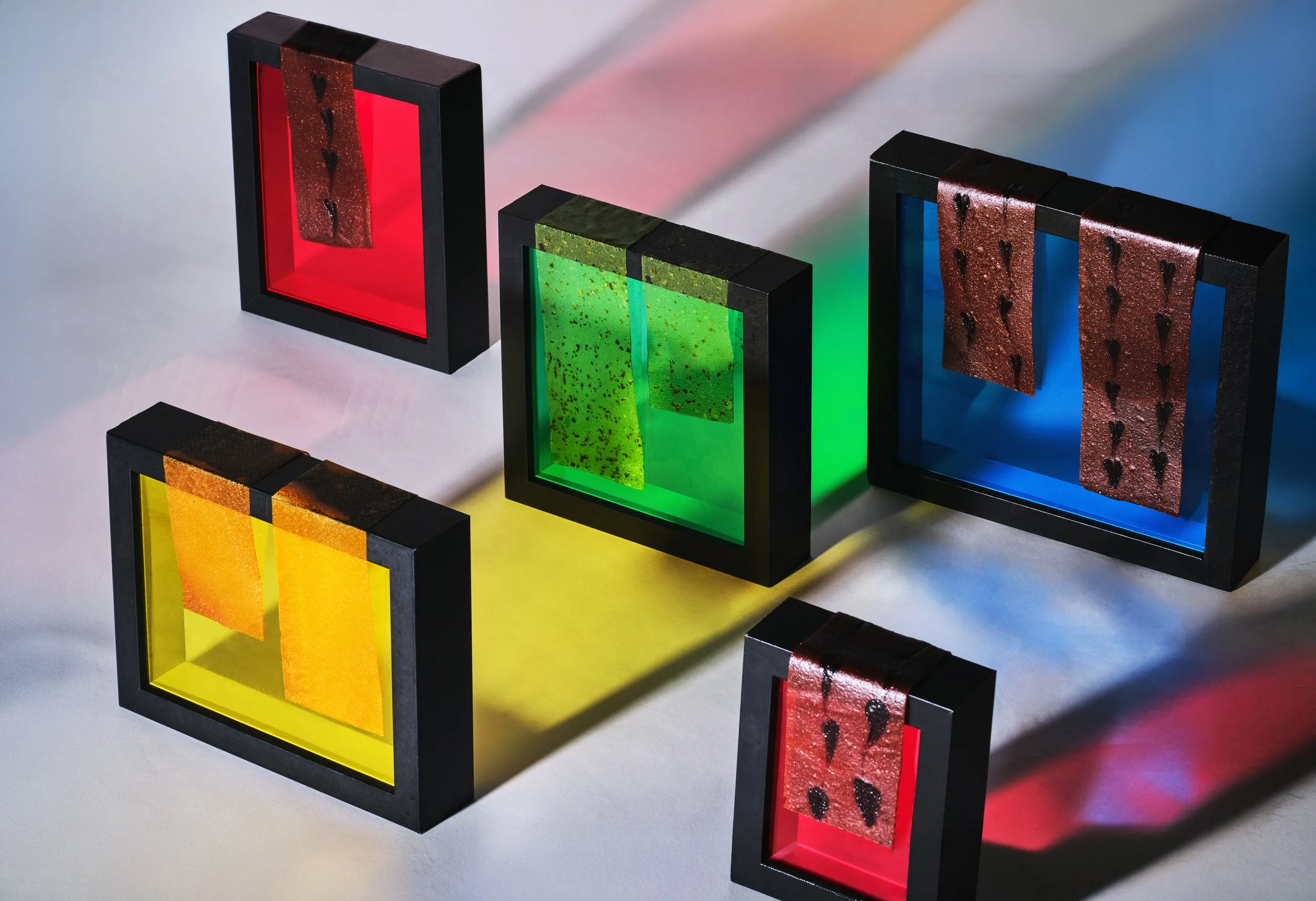 Colorful abstract art pieces in black frames with translucent panels in red, green, blue, and yellow, with liquid or paint-like textures and patterns inside each frame, placed on a white surface with shadows.