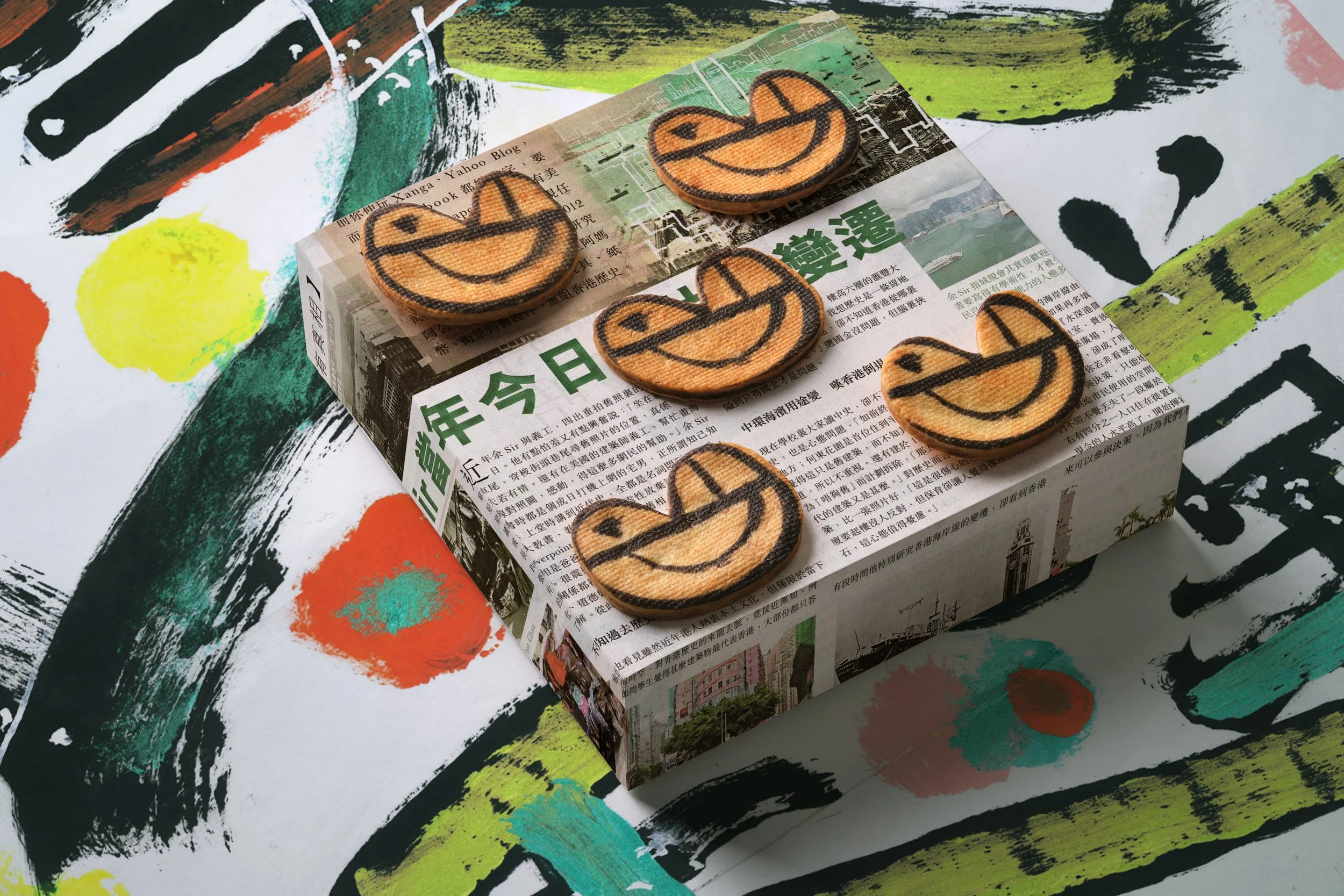 A colorful abstract painted surface with a small newspaper-based box on top. The box has Chinese characters printed on it and six wooden patches with smiling faces outlined in black are placed on top of the box.
