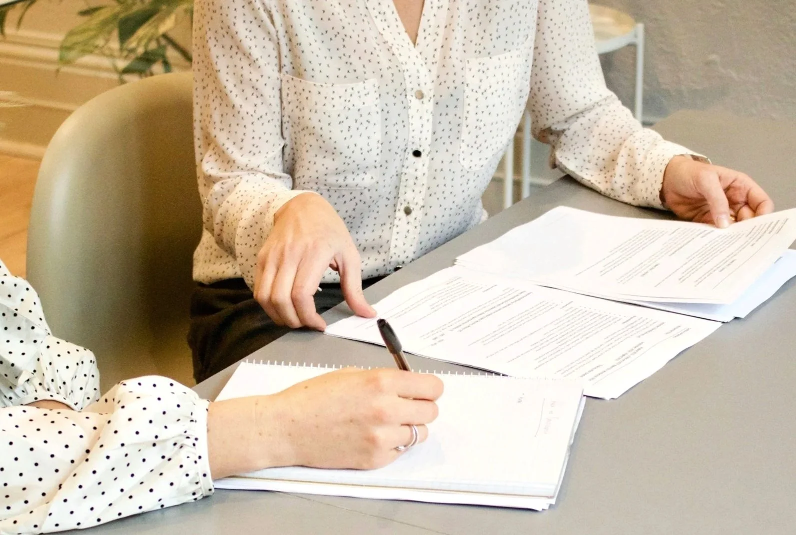 Two people working at a table with stacks of papers, one is writing on a notepad, the other is pointing at documents.