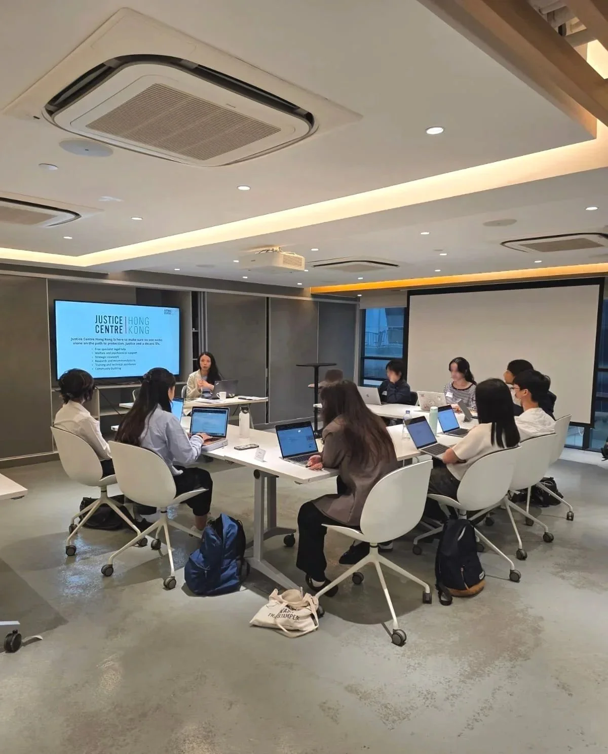 A group of people attending a presentation or meeting in a modern conference room. They are seated around a white table with laptops open, listening to a woman speaker. There is a large screen displaying information about the Justice Centre Hong Kong.