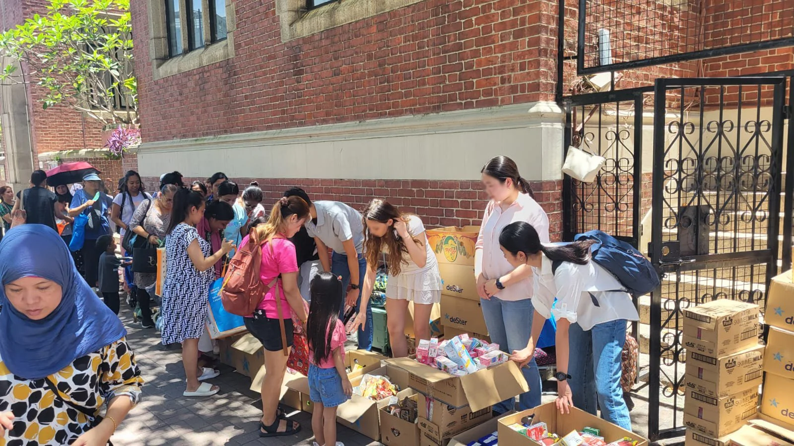 People lining up at a charity food distribution event outside a brick building, receiving free food items such as milk, snacks, and canned goods.