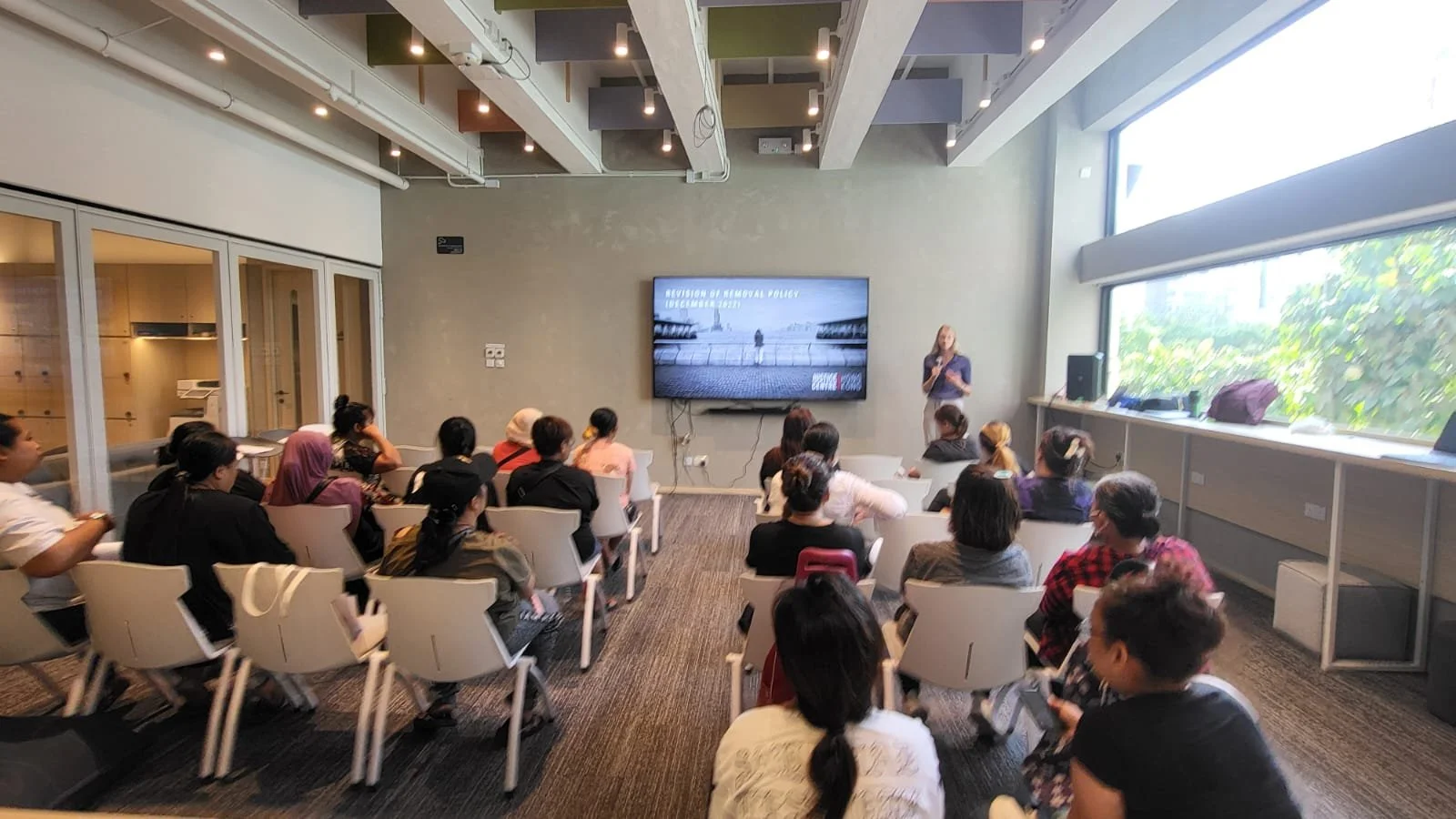 A group of people attending a presentation in a modern conference room with a large window; the presenter is standing near a screen displaying a slide about the revision of the removal policy.