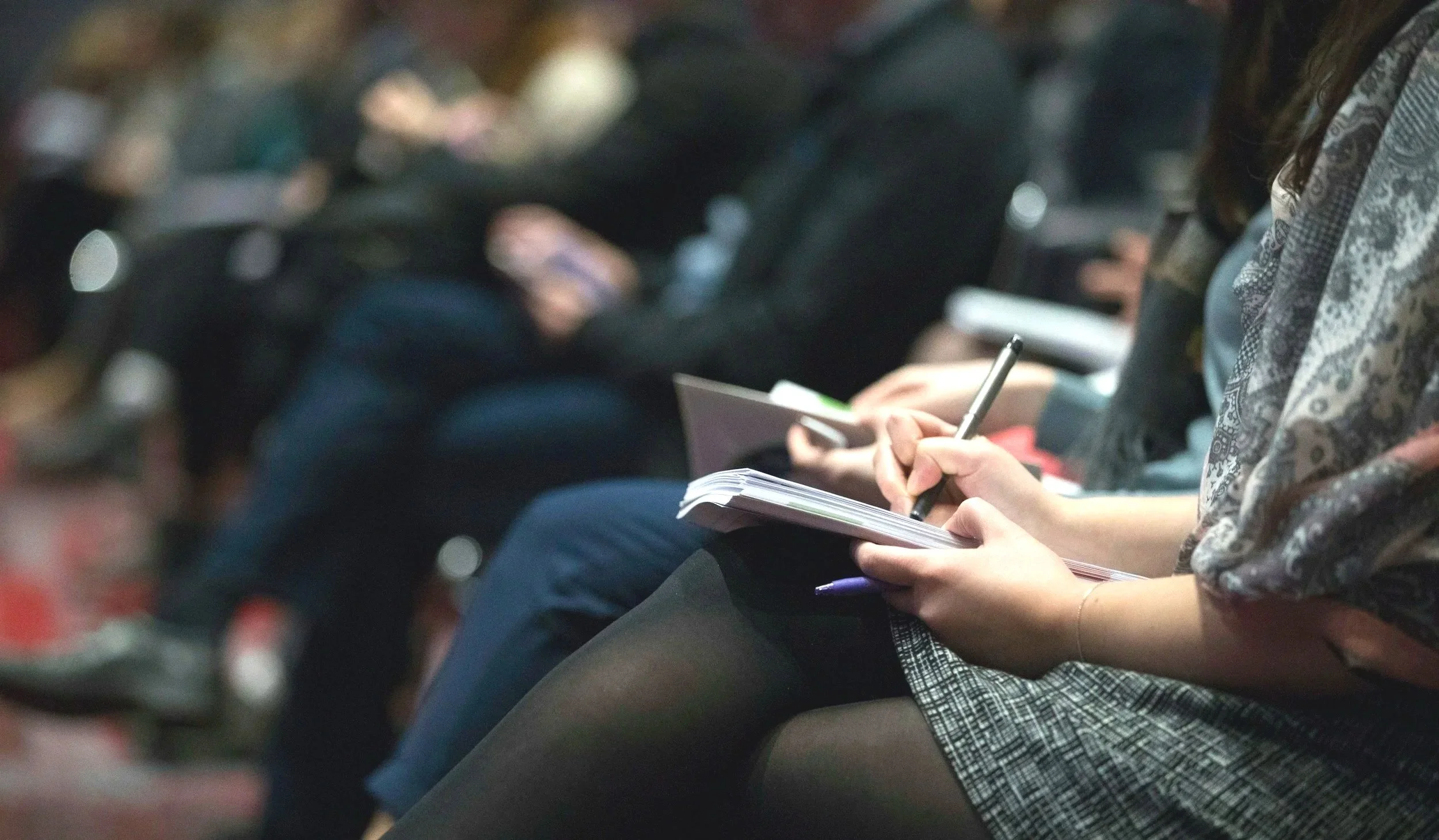Close-up of a person sitting in an audience, taking notes with a pen on a notepad, wearing a patterned scarf or shawl.