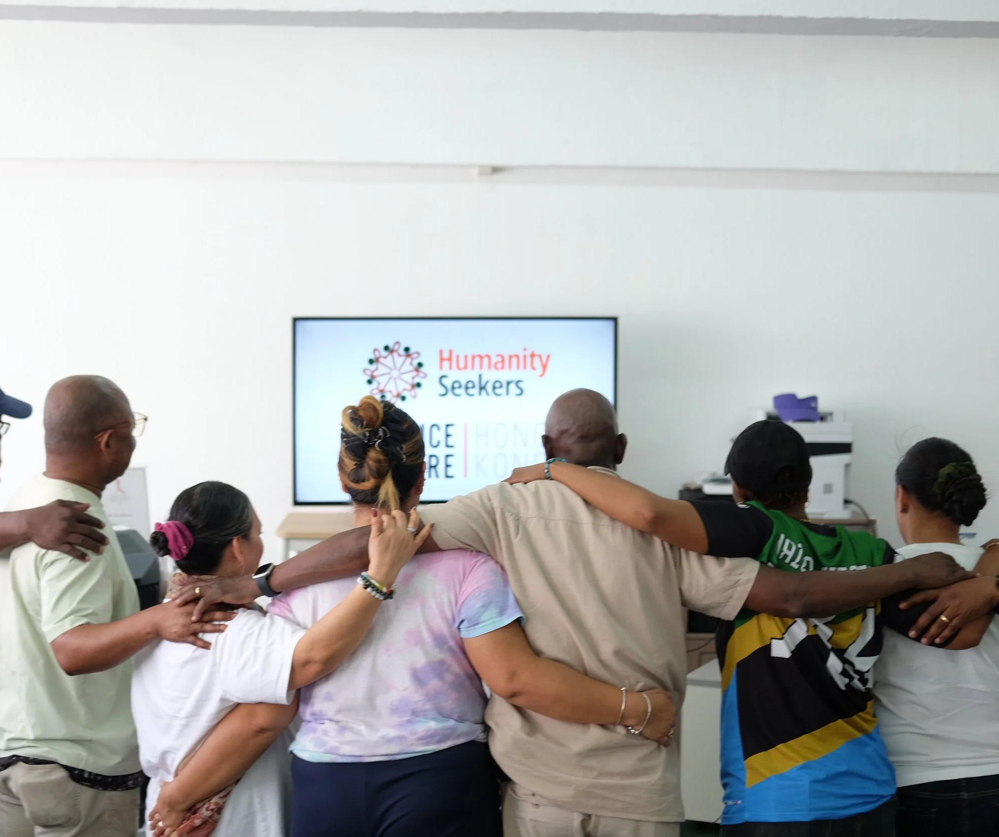 A group of diverse people in a supportive hug, standing in front of a screen displaying the Humanity Seekers logo at a gathering or event.