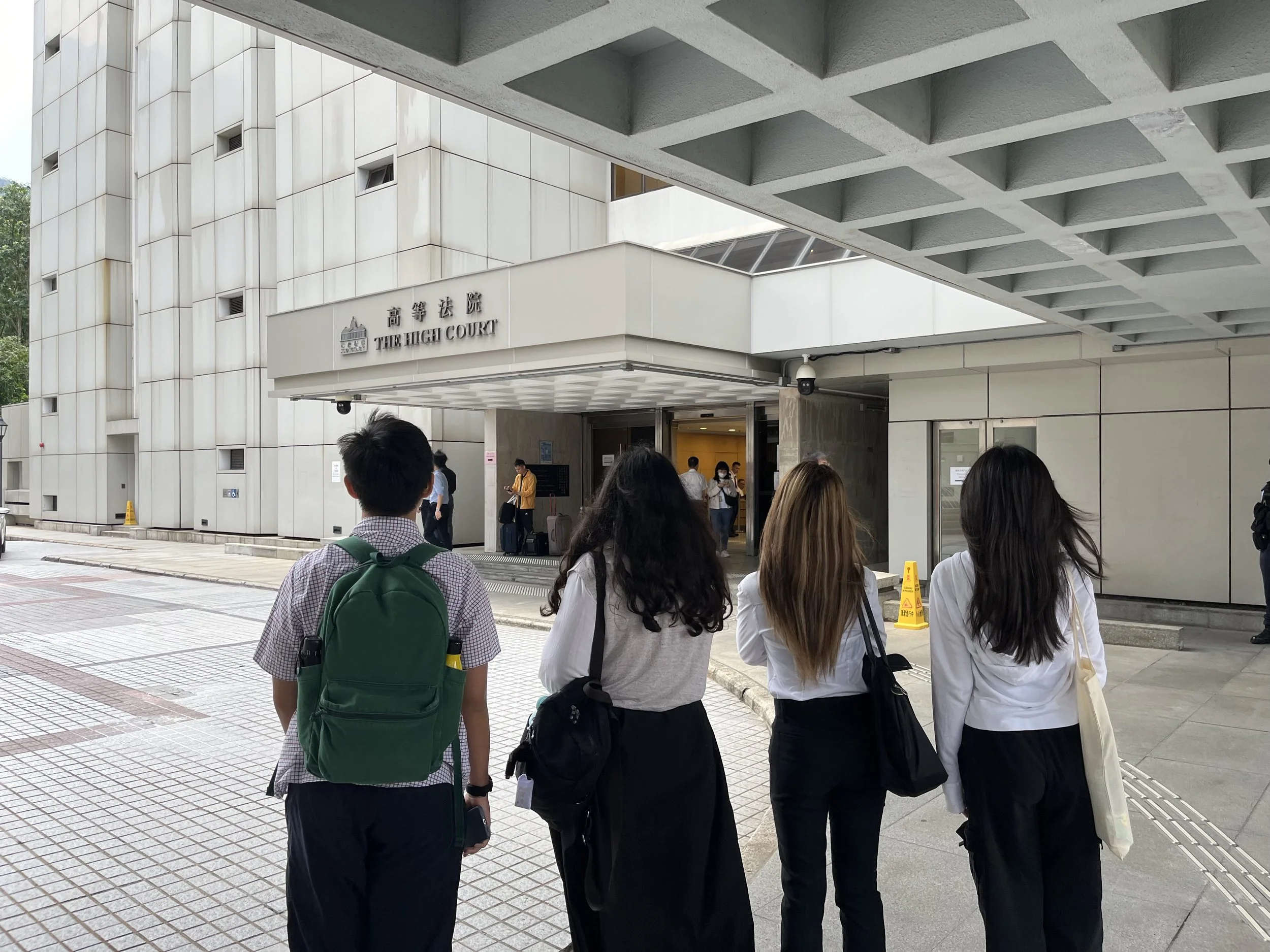 People waiting outside of the High Court building in daytime, some with luggage and backpacks, under a covered area with a modern architectural design.