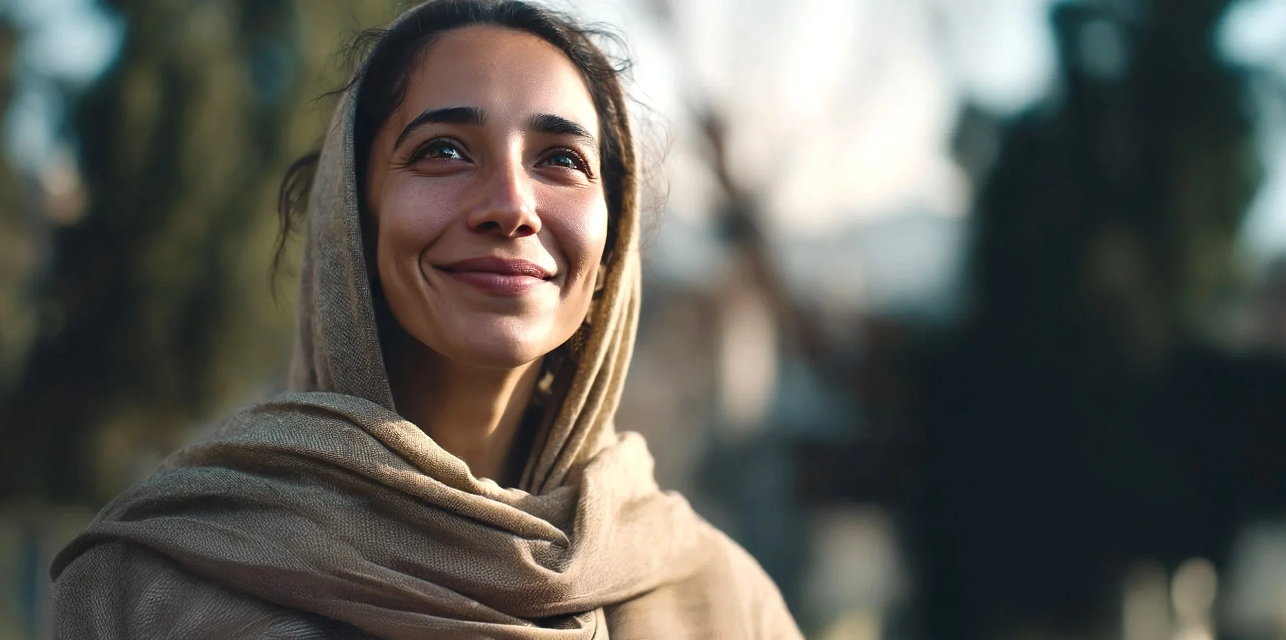 A woman with dark hair and brown eyes smiling outdoors, wearing a beige hoodie with the hood over her head.