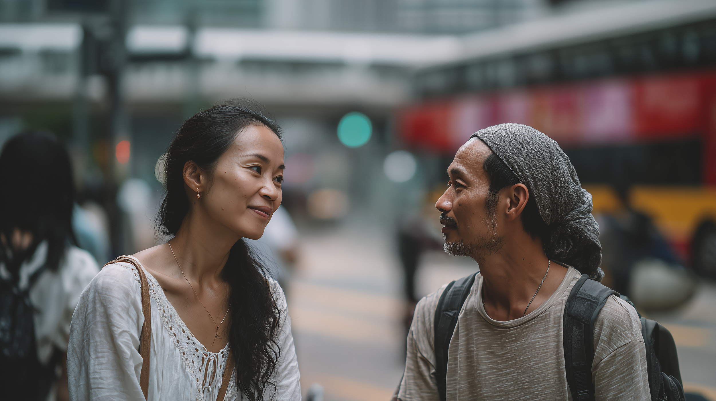 A woman with long black hair and a man with a gray bandana talking outdoors in an urban setting, with blurred city background and other people around.