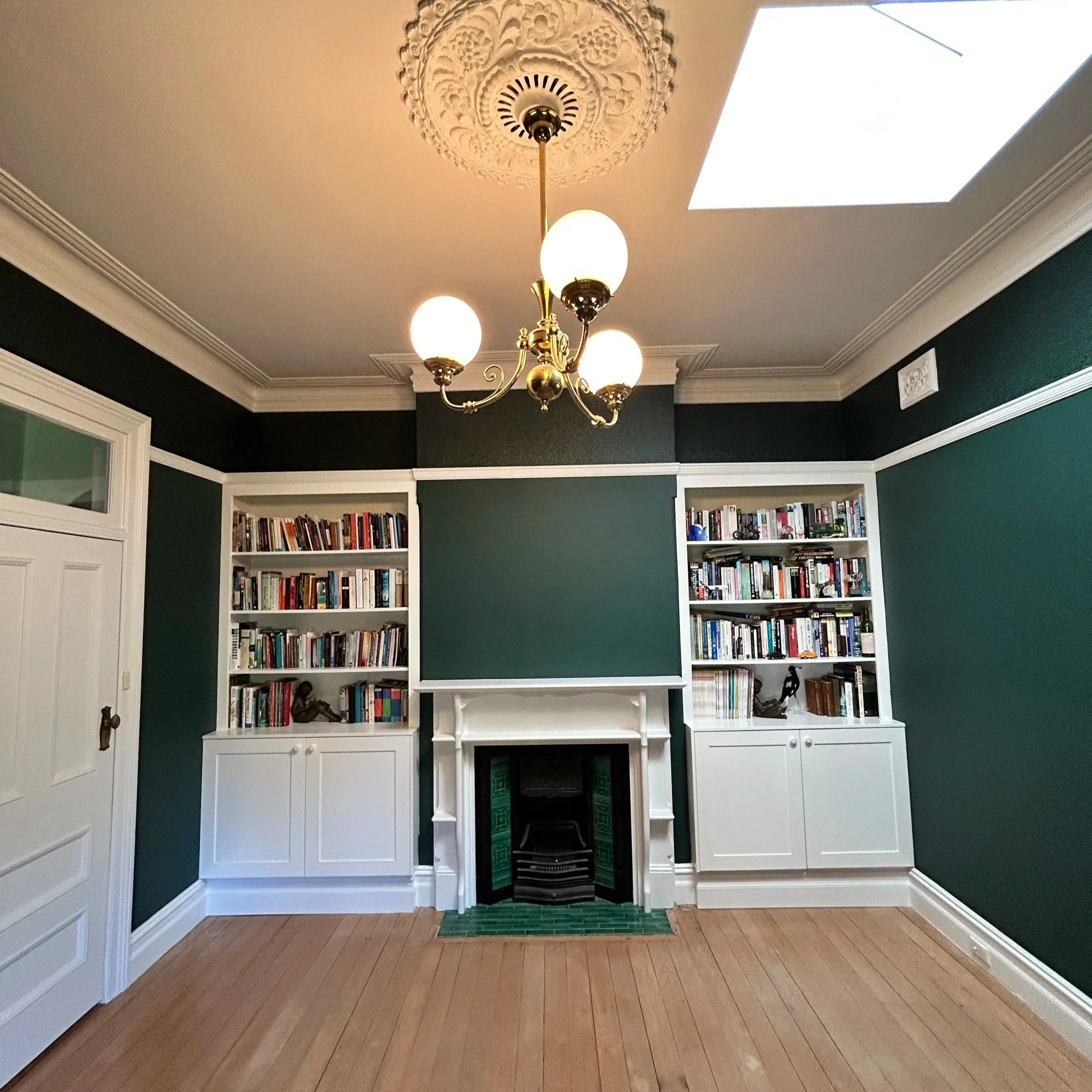 A room with dark green walls, white bookshelves filled with books on each side of a white fireplace with green tiles inside, a wooden floor, and a brass chandelier with five globes hanging from ornate ceiling molding. A skylight is visible in the cei