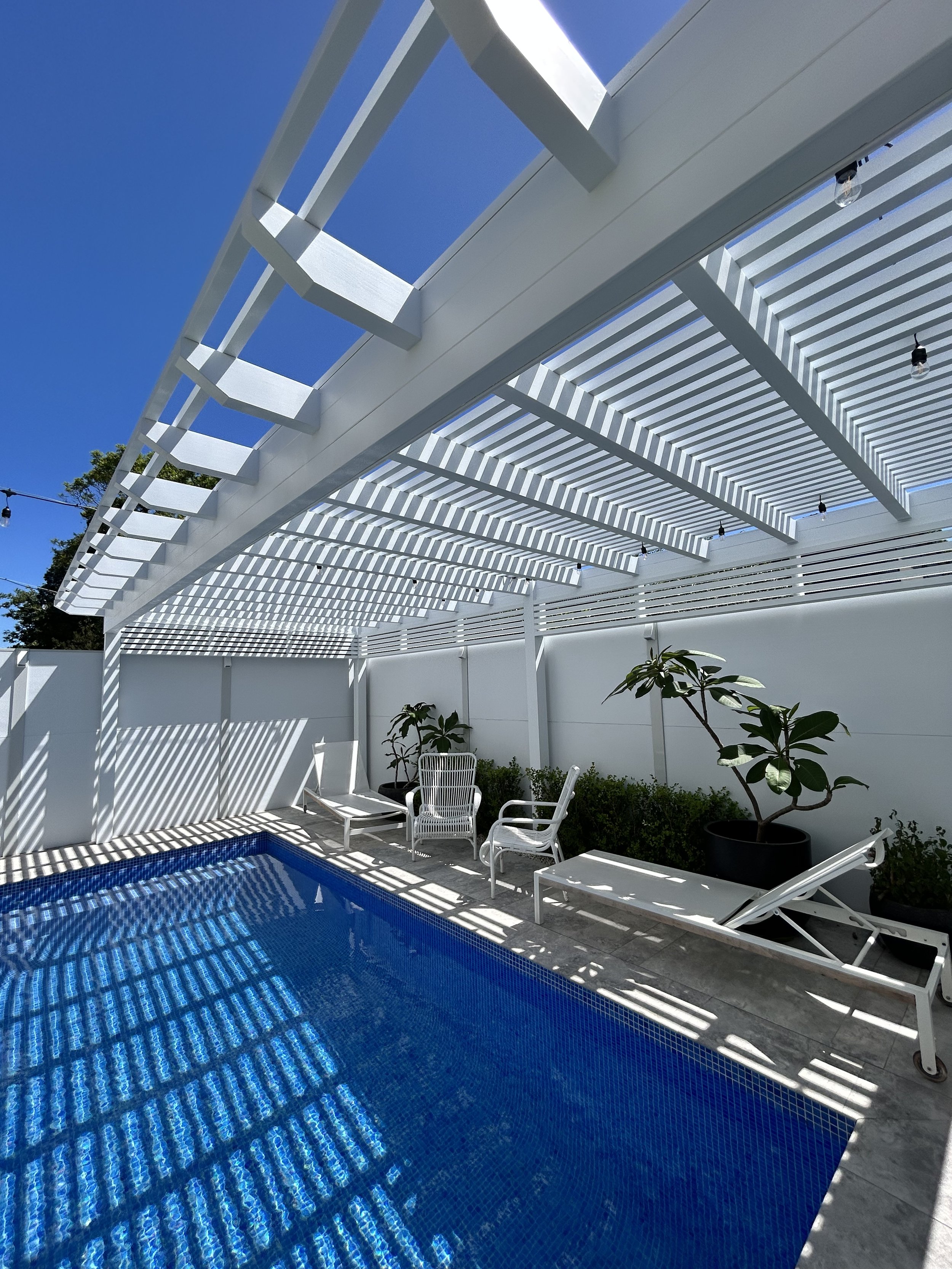 Backyard pool with white lattice pergola casting striped shadows on the pool and patio furniture, including white lounge chairs and potted plants, under a clear blue sky.