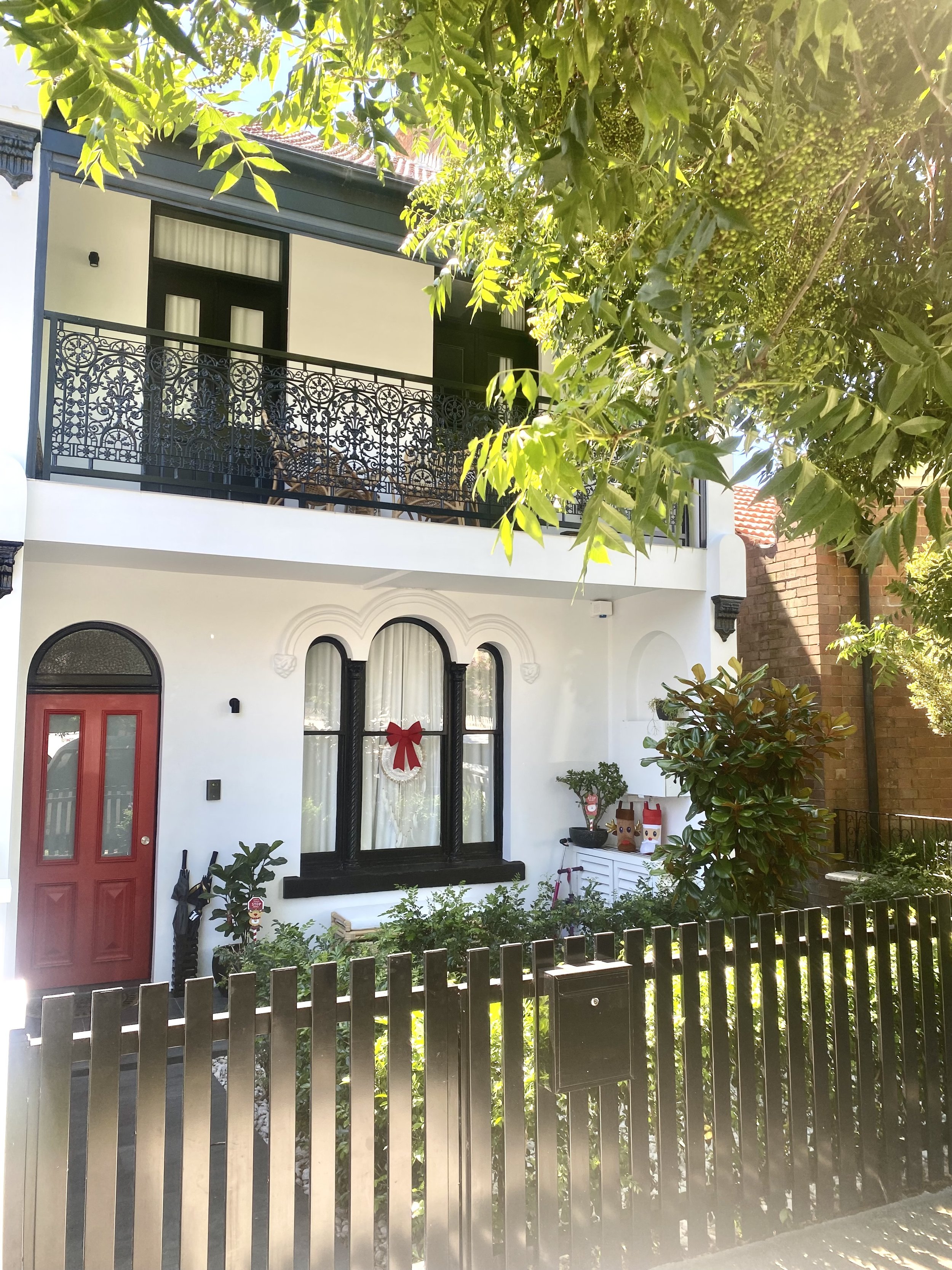 Front view of a white house decorated for Christmas with a red bow on a window, holiday figures on a white table, and a garden, surrounded by a black picket fence and shaded by trees.