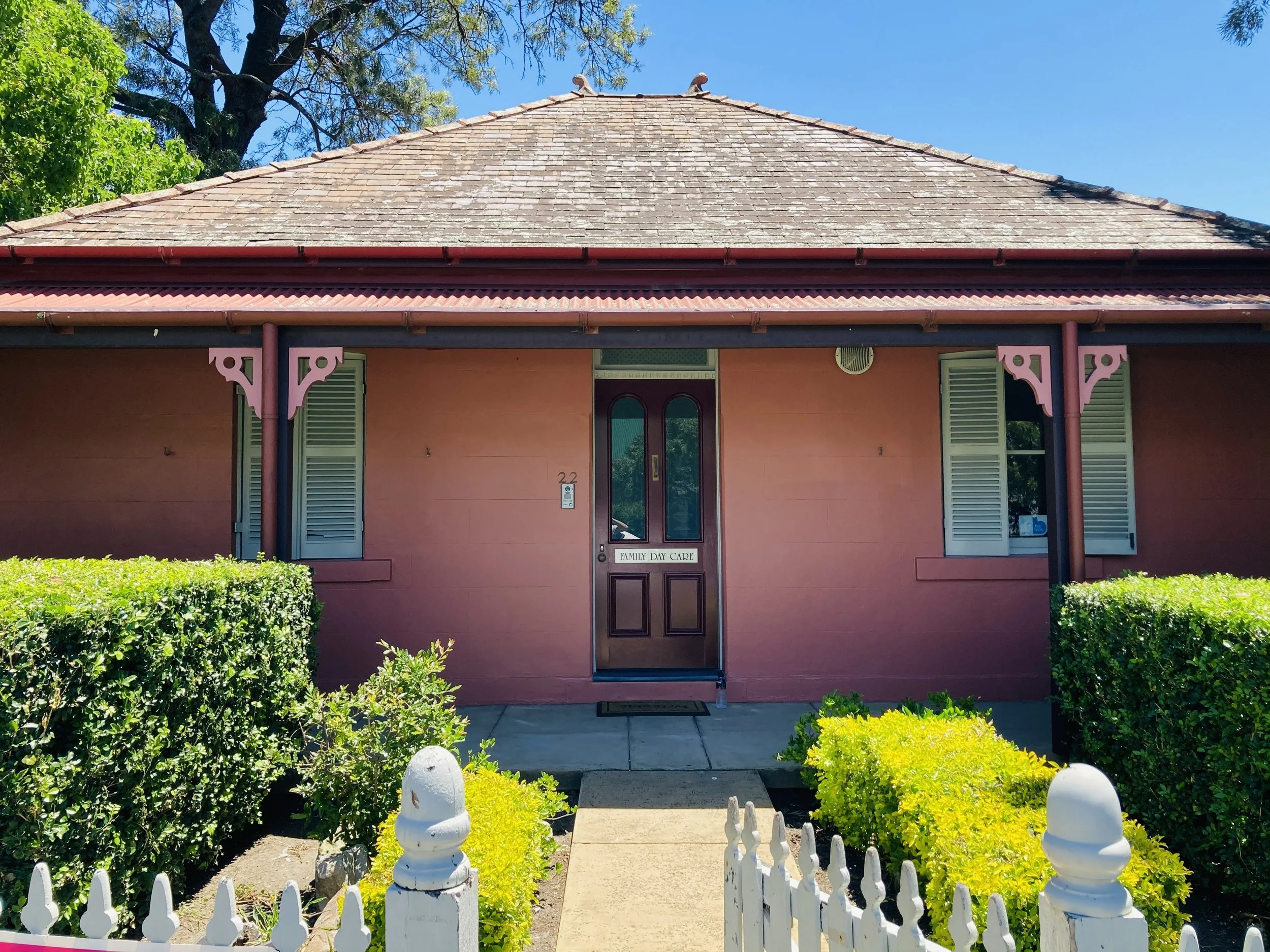 Pink house with a brown door labeled 'Family Day Care', green bushes, white picket fence, and a decorative porch with pink architectural elements under a clear blue sky.