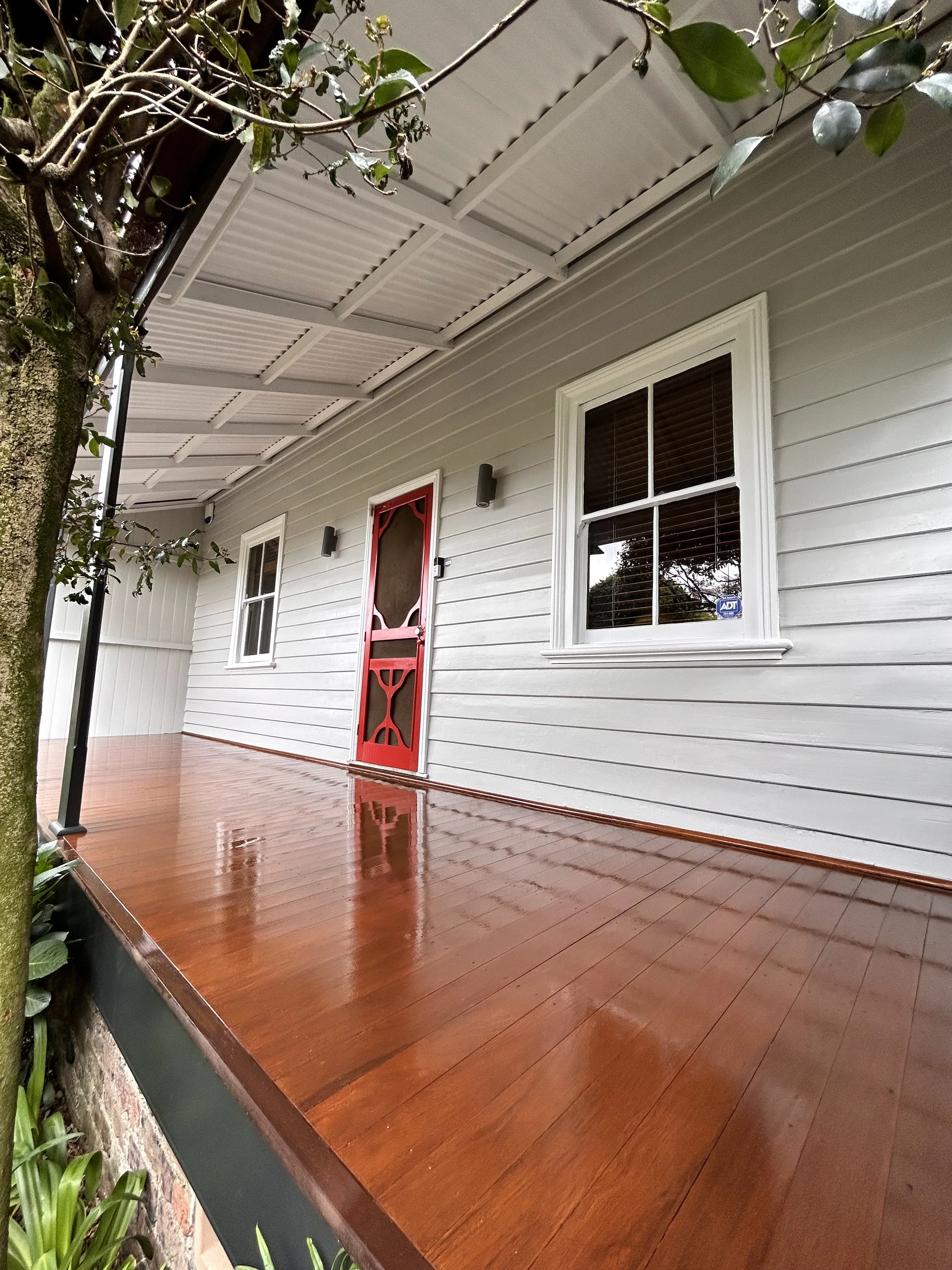 Front porch of a house with a polished wooden floor, white siding, two white-framed windows, and a red door, surrounded by greenery.