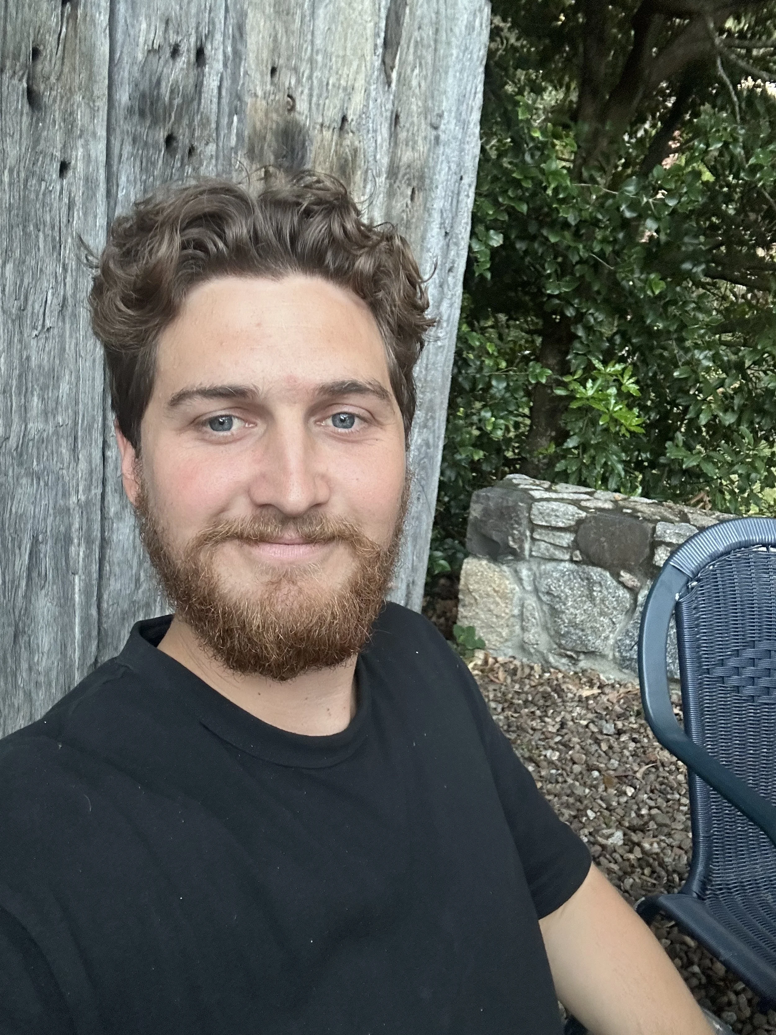 A person with curly hair and a beard smiling at the camera, sitting outdoors near a wooden surface and foliage, with a partial view of a chair.