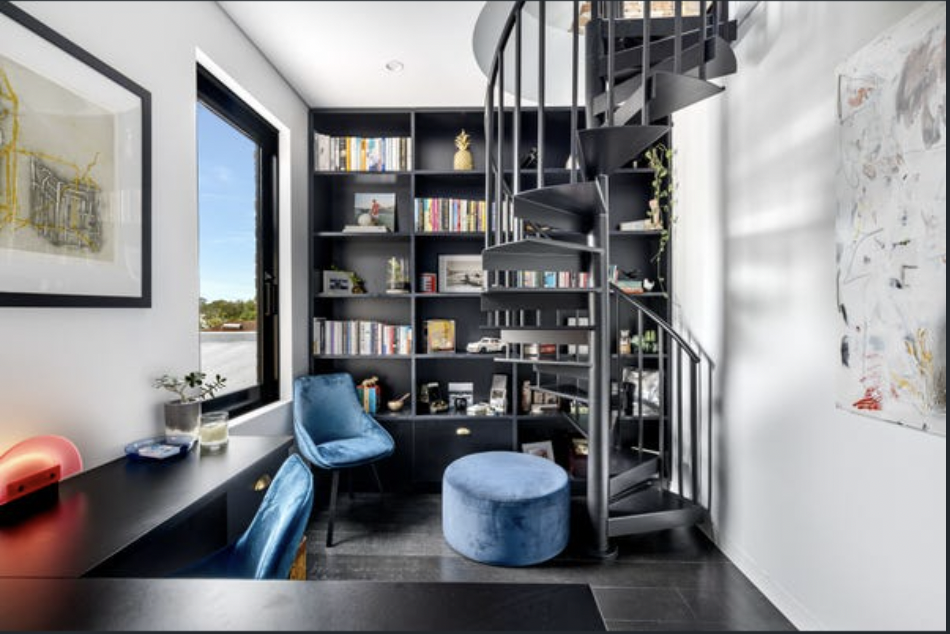 Modern home office with a black spiral staircase, bookshelves, blue velvet chairs, desk, and abstract artwork on the wall.