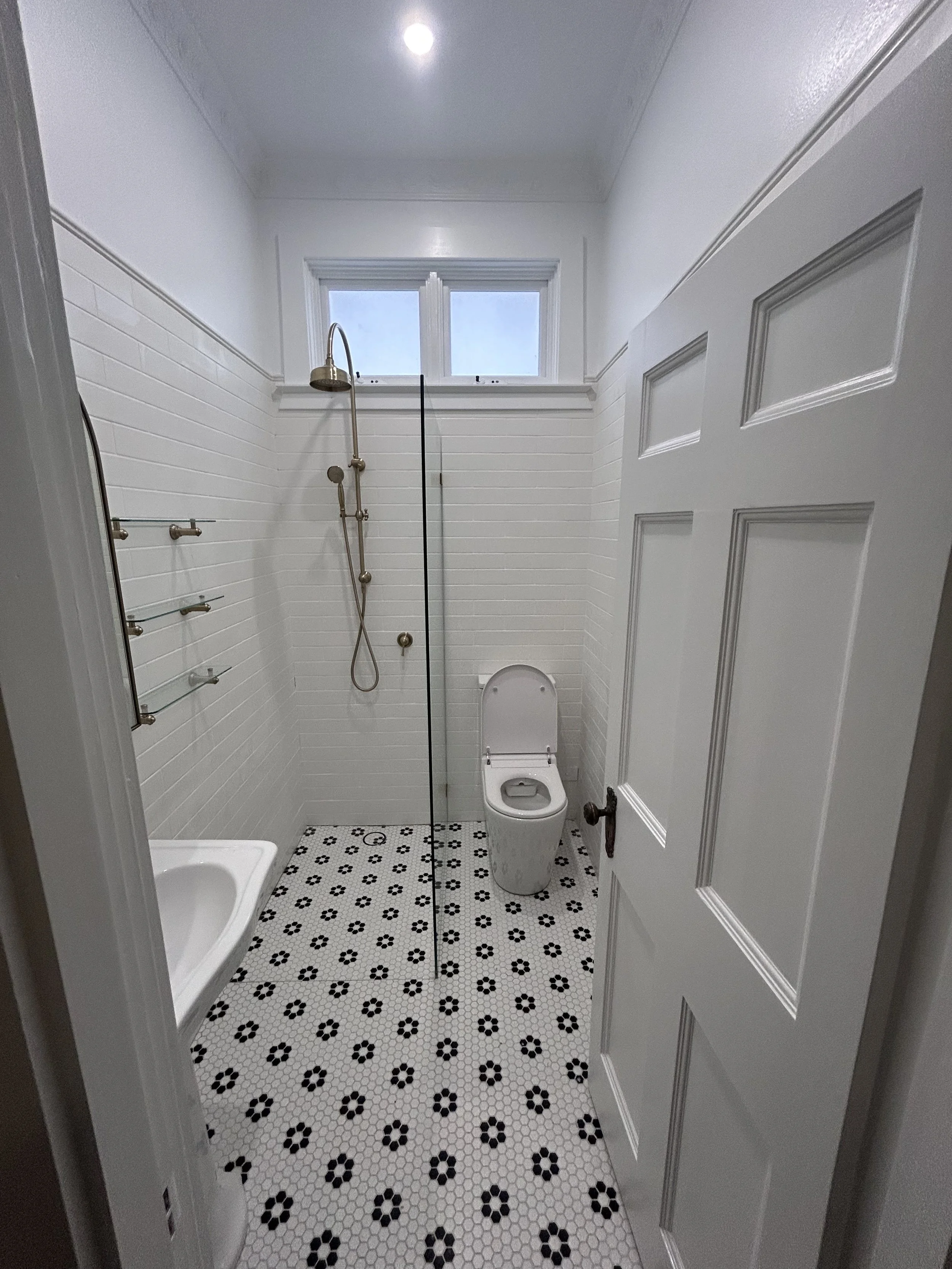 Small bathroom with white tiled walls, a window above, a showerhead on the wall, a toilet, a small sink, a glass partition, and black and white patterned hexagonal floor tiles.