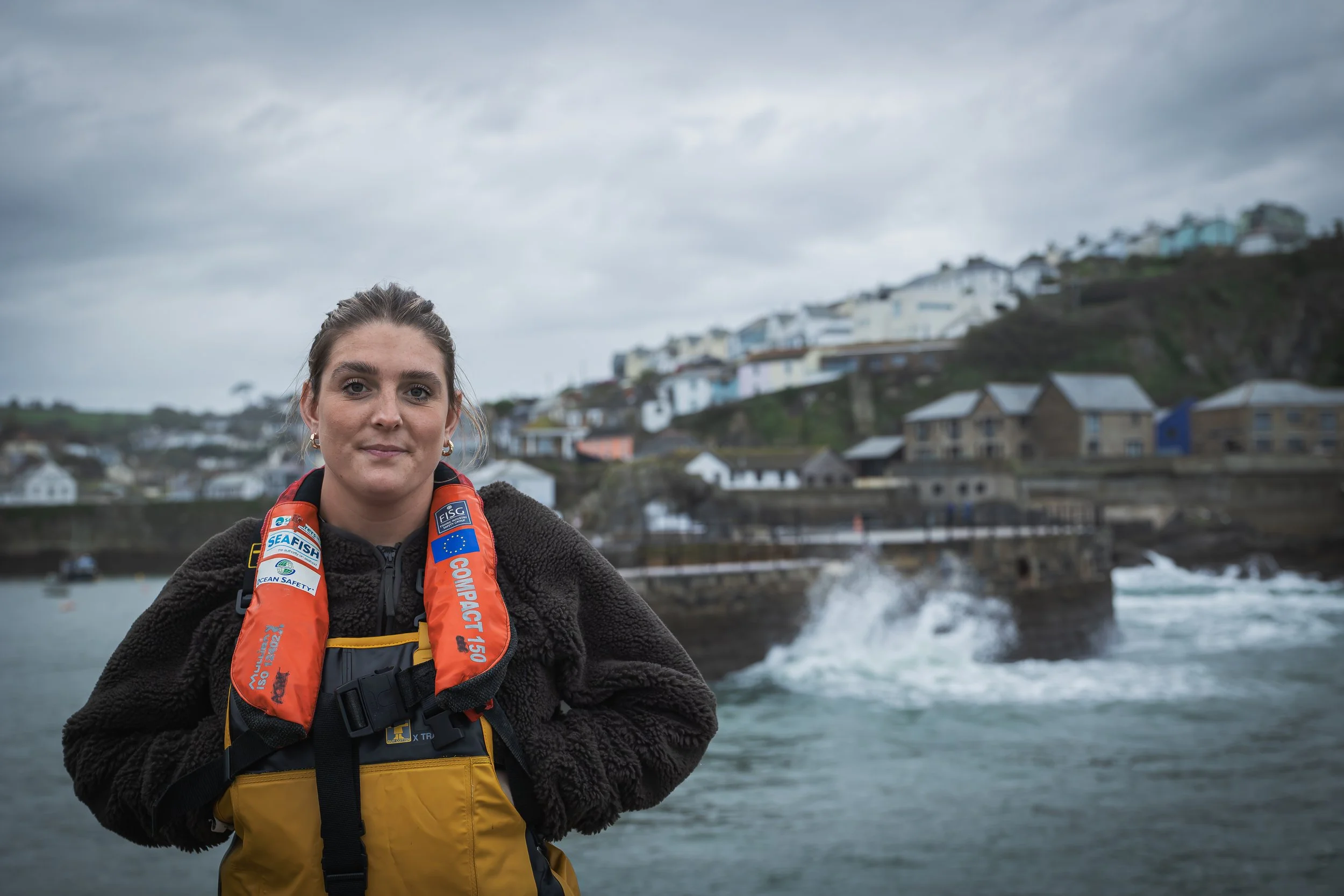 A young woman in a PFD and oilskins above and in front of a harbor being battered by waves