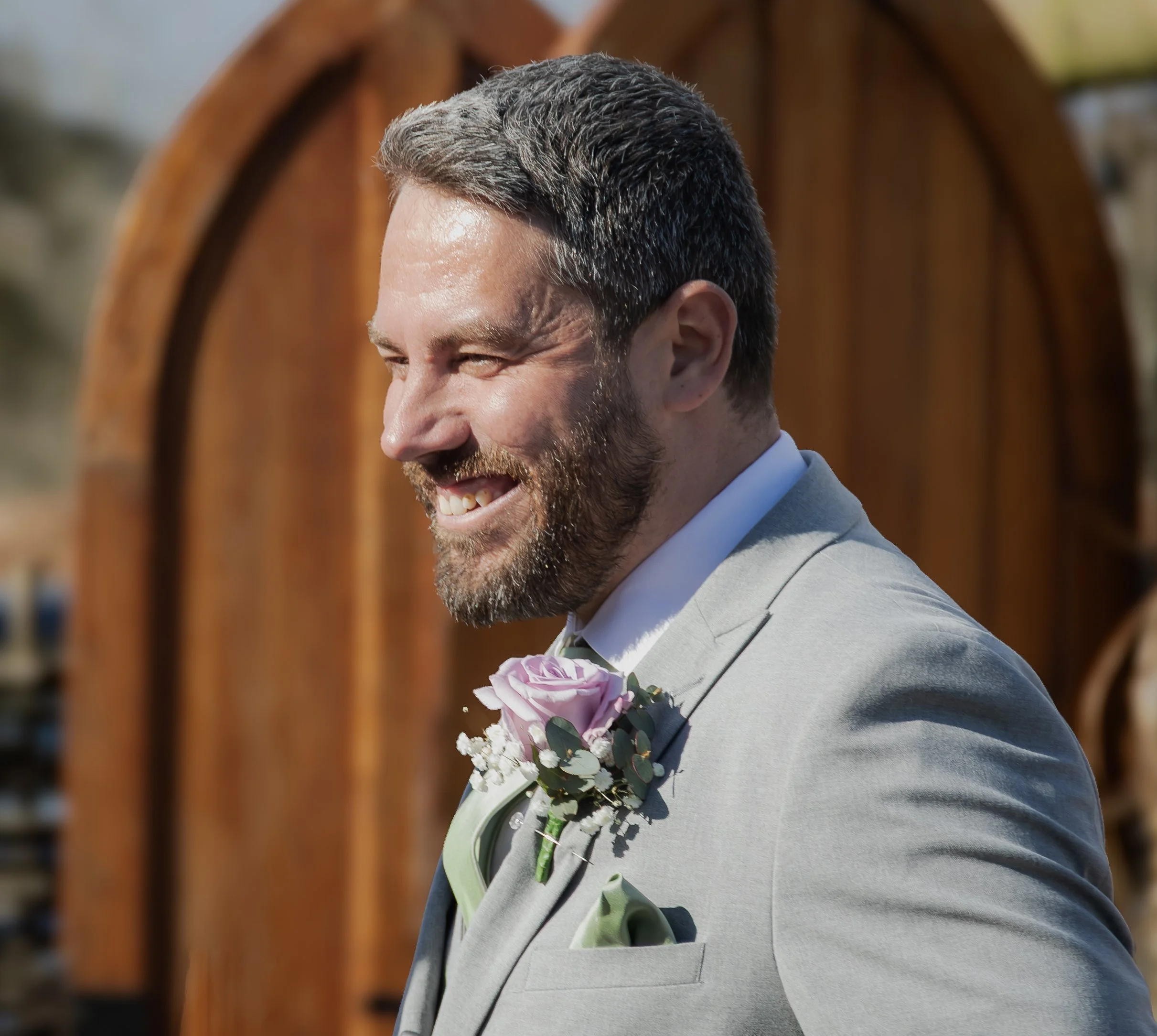 A smiling man at a wedding in a grey suit with a flower
