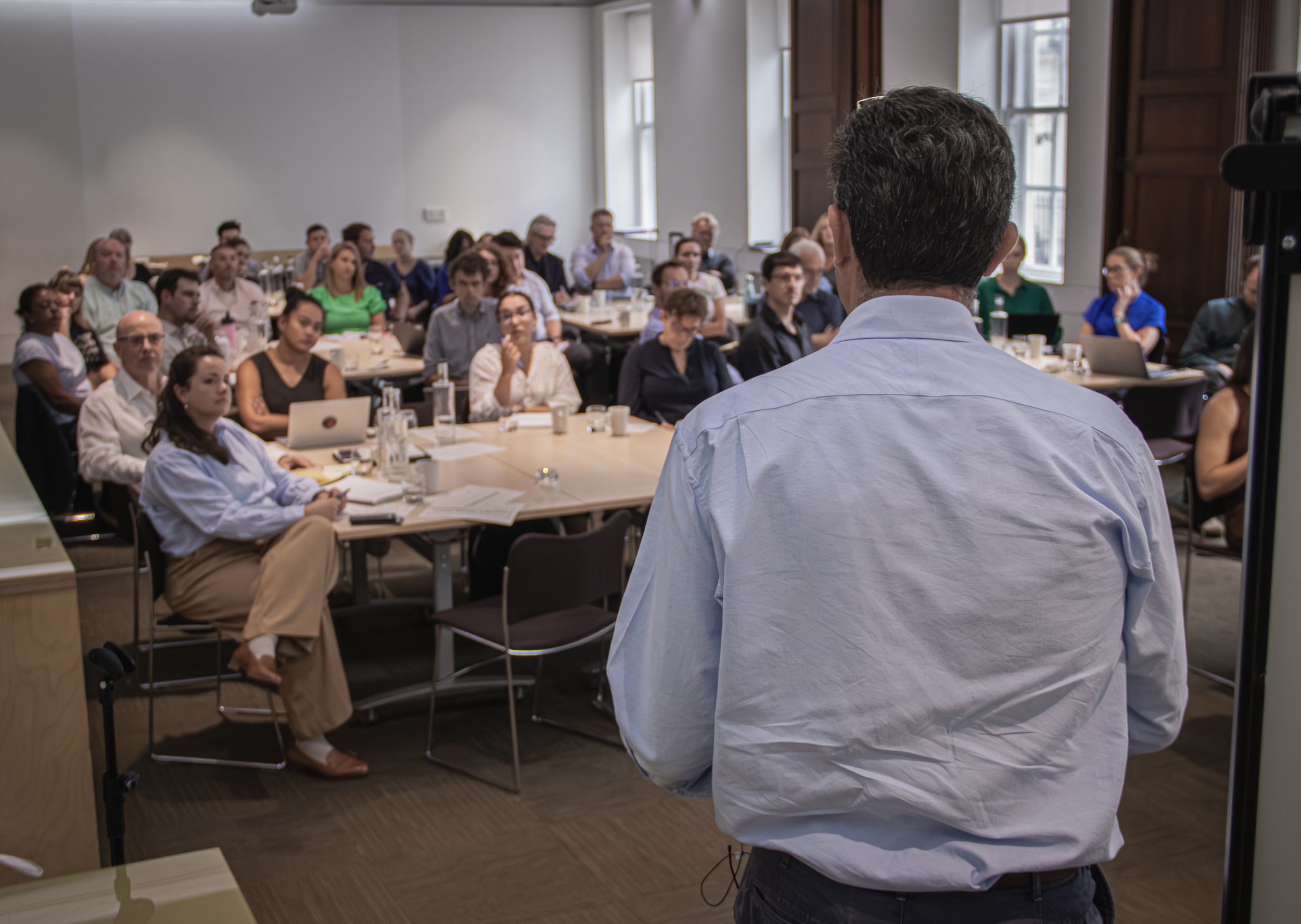An image from behind a man giving a presentation, speaking to a large crowd of people in chairs