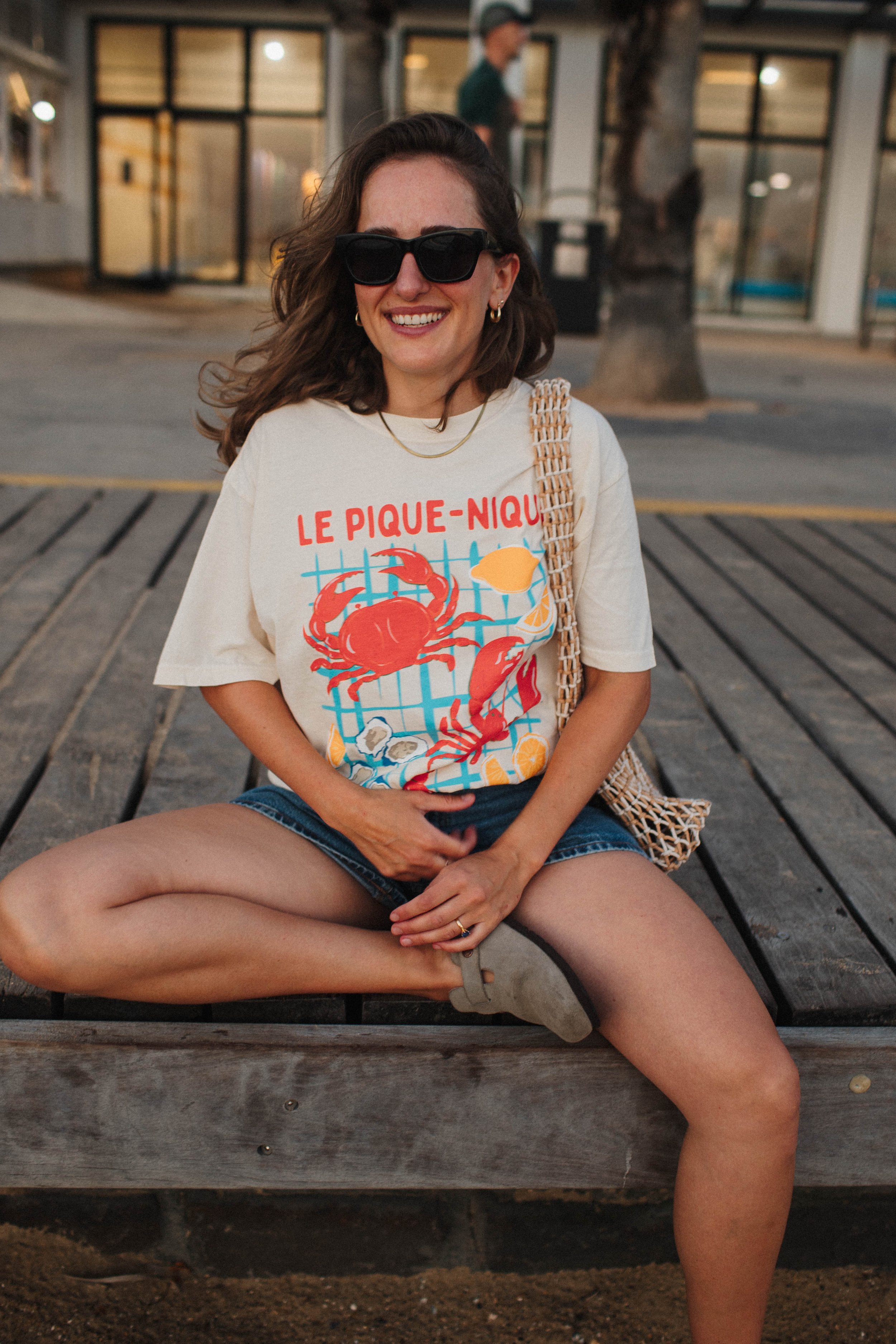 A woman sitting on a wooden bench, wearing sunglasses, a shirt with "Le Pique-Nique" and a crab design, denim shorts, and holding a mesh bag.