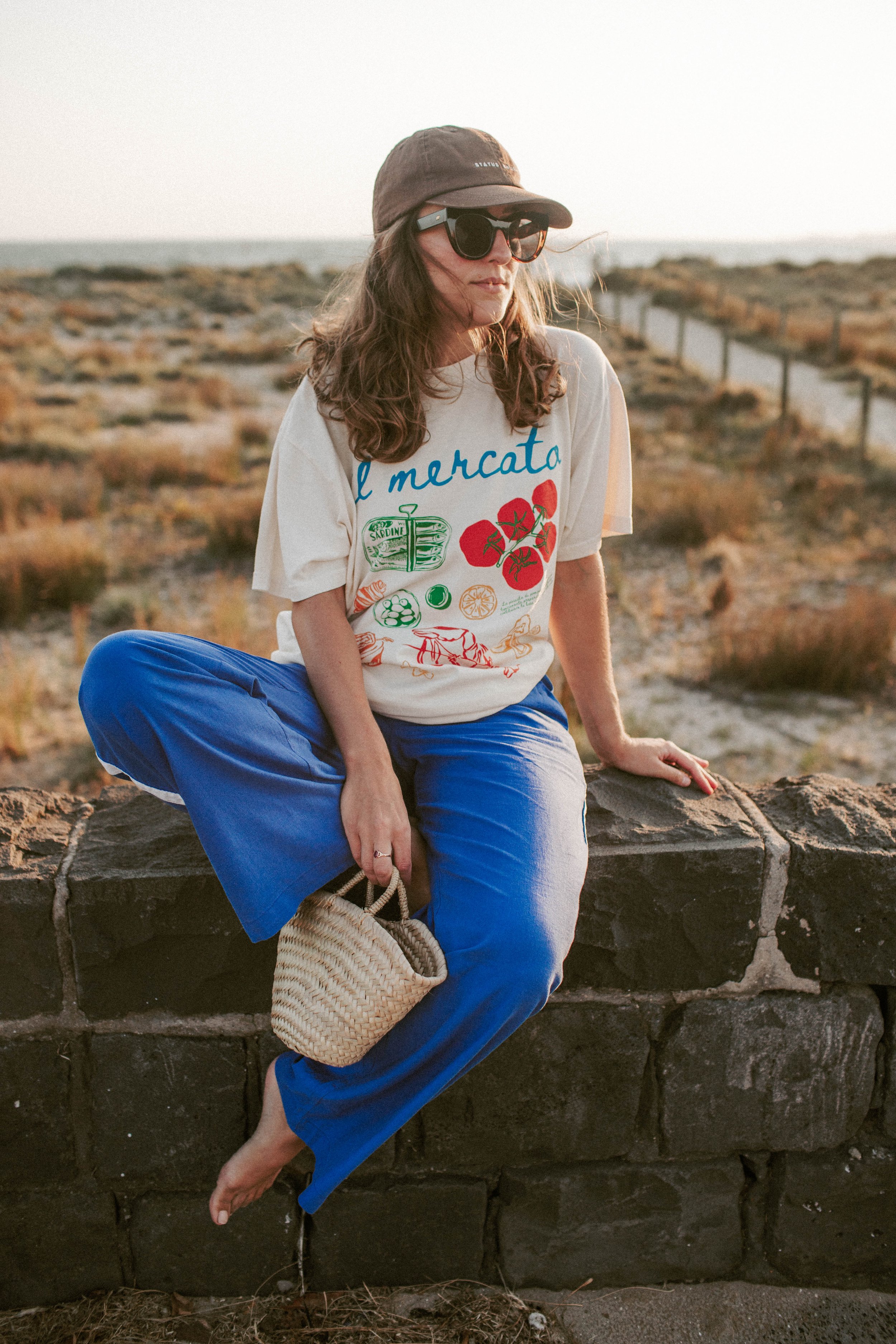 Person wearing sunglasses and a cap, sitting on a stone wall in outdoor setting. They are dressed in a graphic t-shirt with 'Il Mercato' text and illustrations of food, blue pants, holding a woven bag with a natural landscape in the background.