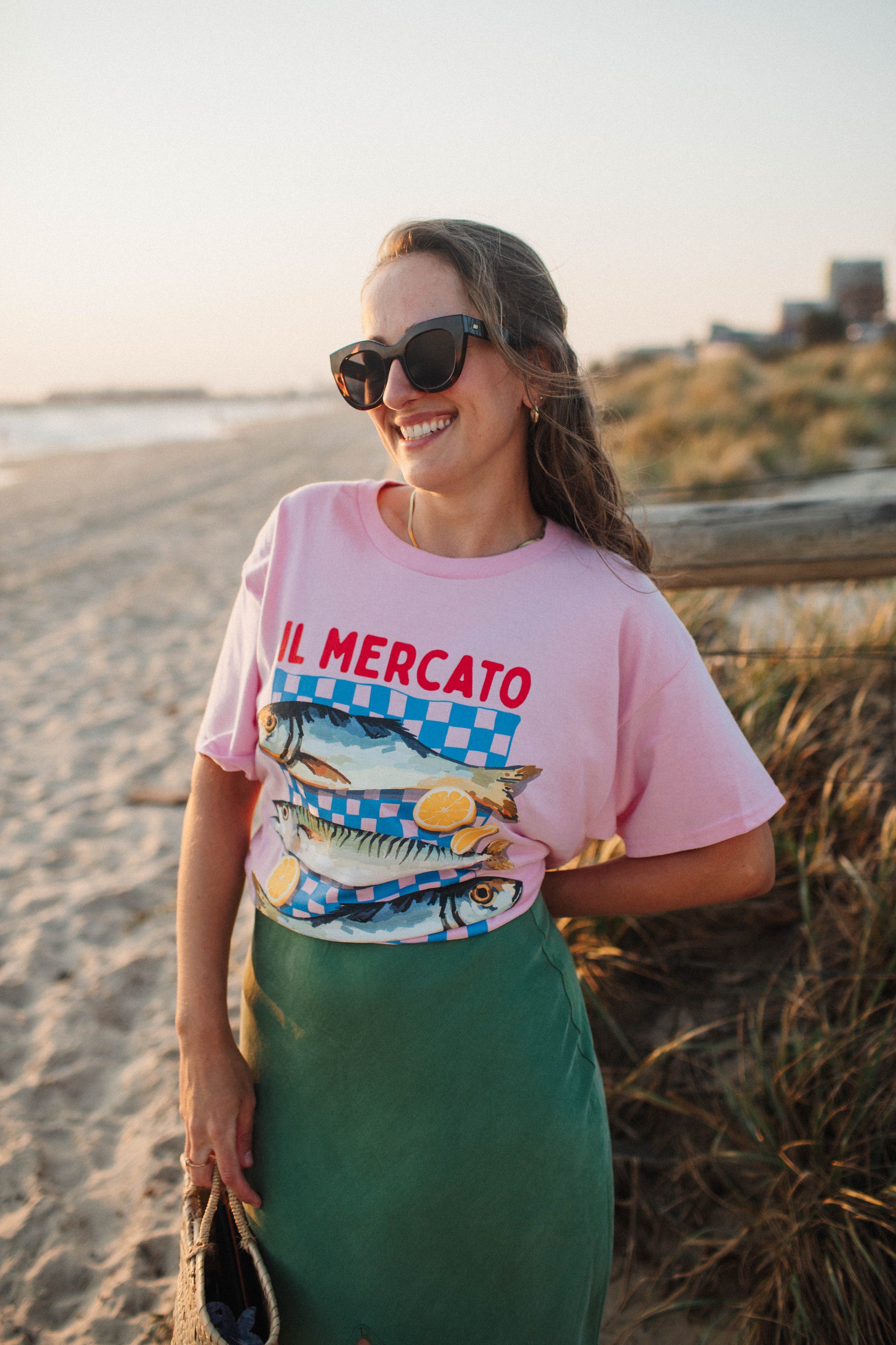 Woman wearing sunglasses and a pink "IL MERCATO" t-shirt with fish design, standing on a beach.