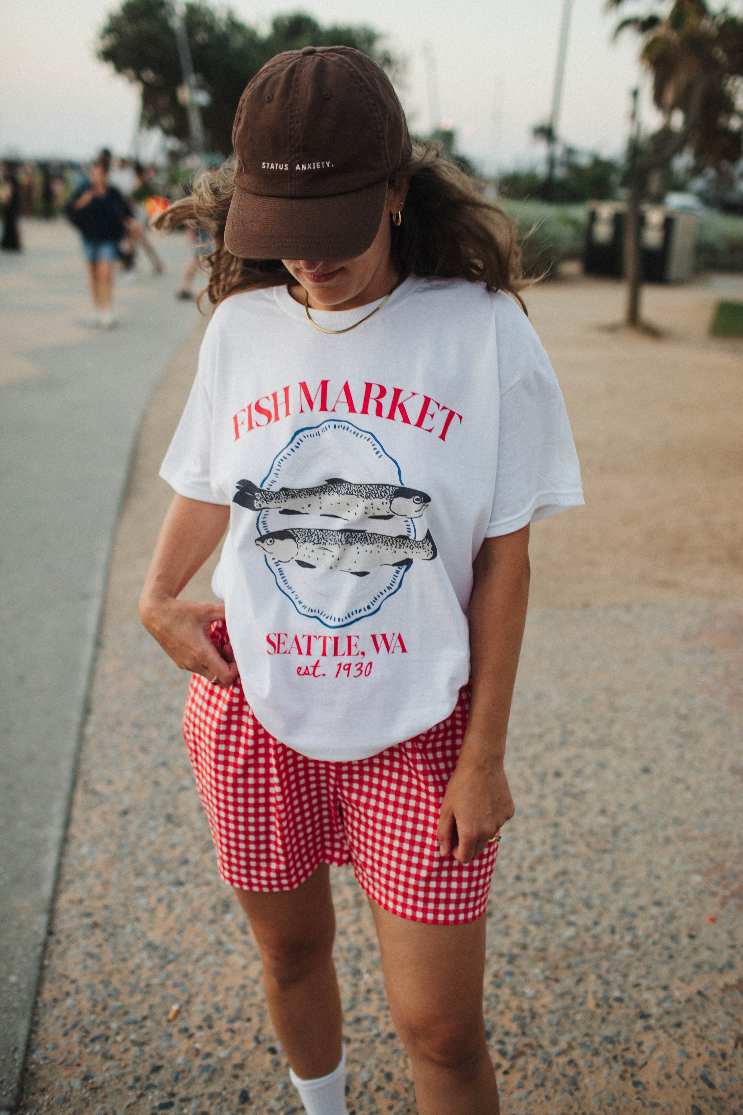 Person wearing a white t-shirt with "Fish Market Seattle, WA" print and red checkered shorts, standing outdoors.