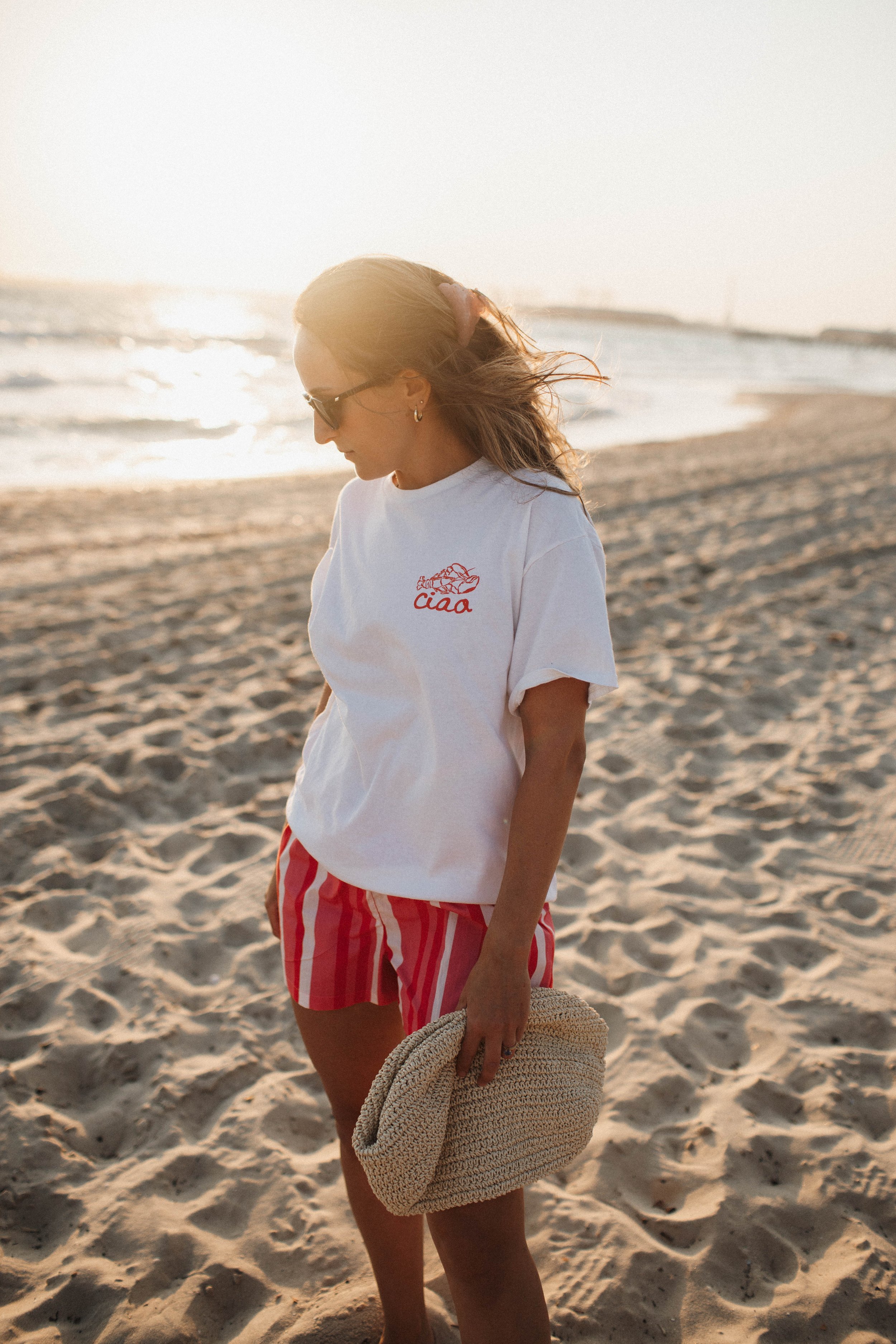 A woman wearing sunglasses, a white T-shirt with "ciao" printed on it, and red striped shorts stands on a sandy beach, holding a woven bag, with the sun setting over the ocean in the background.