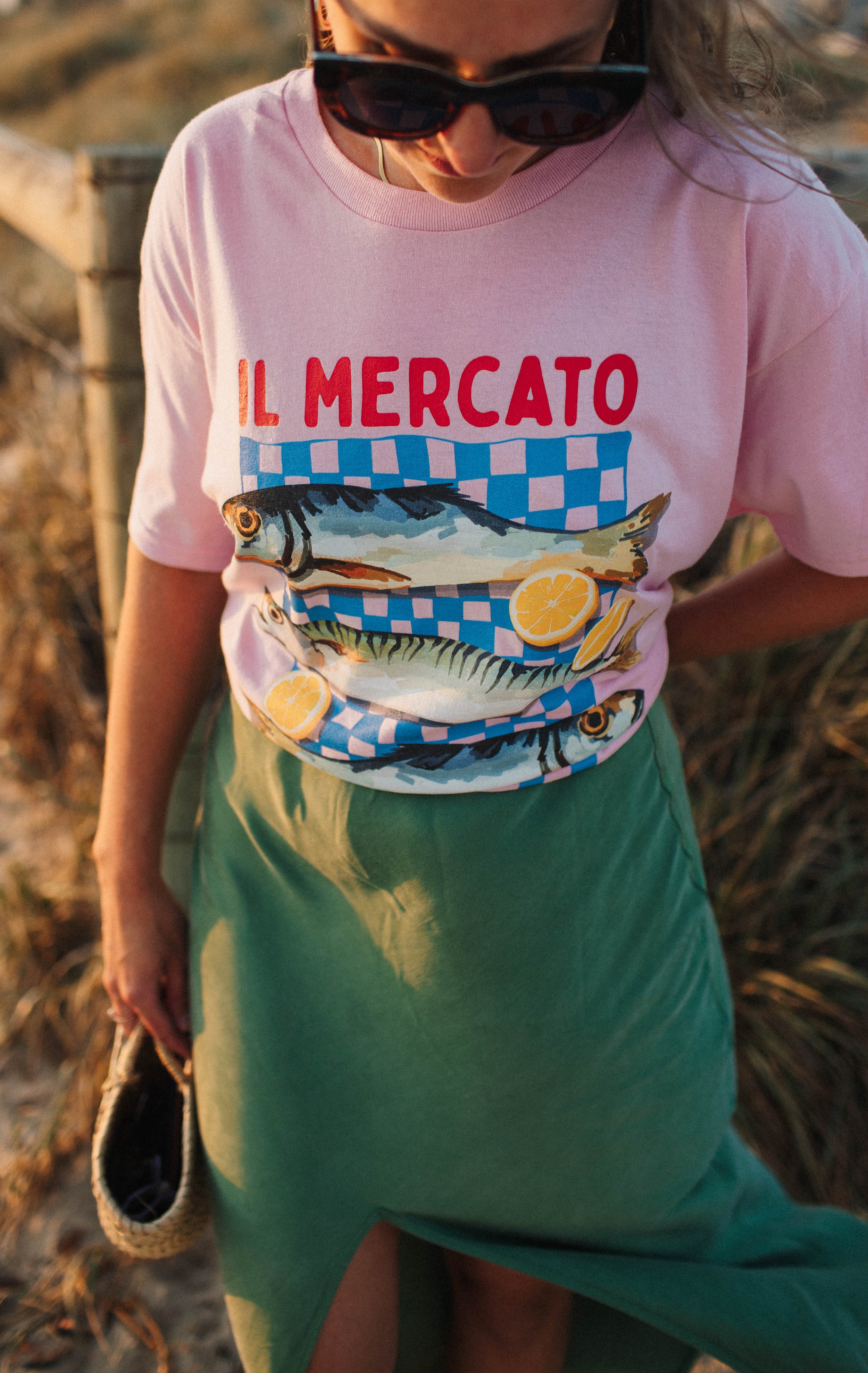 Person wearing a pink shirt with "IL MERCATO" text and fish design, holding a straw handbag, standing outdoors.