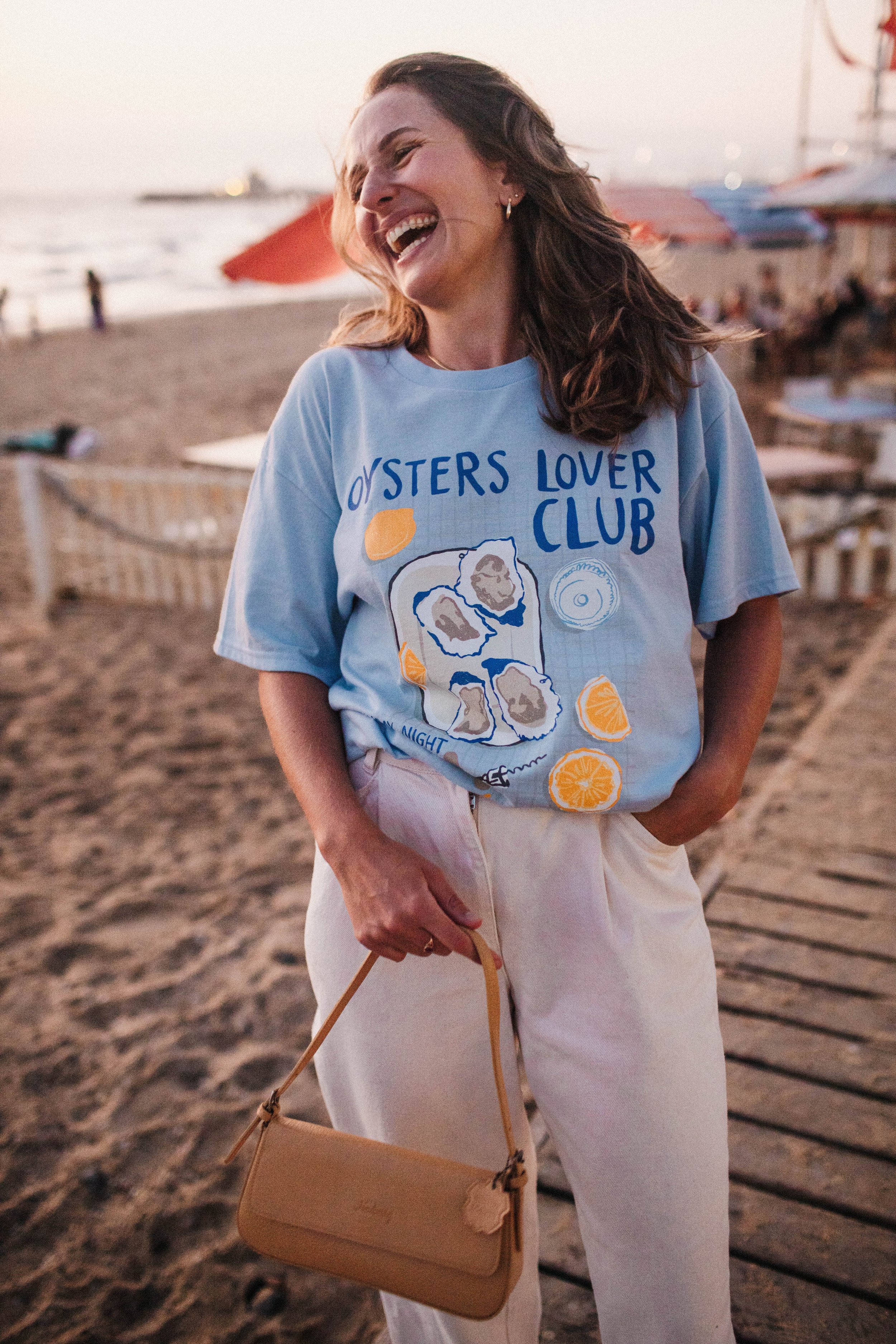 Woman on beach wearing 'Oysters Lover Club' shirt, holding a tan purse, smiling.
