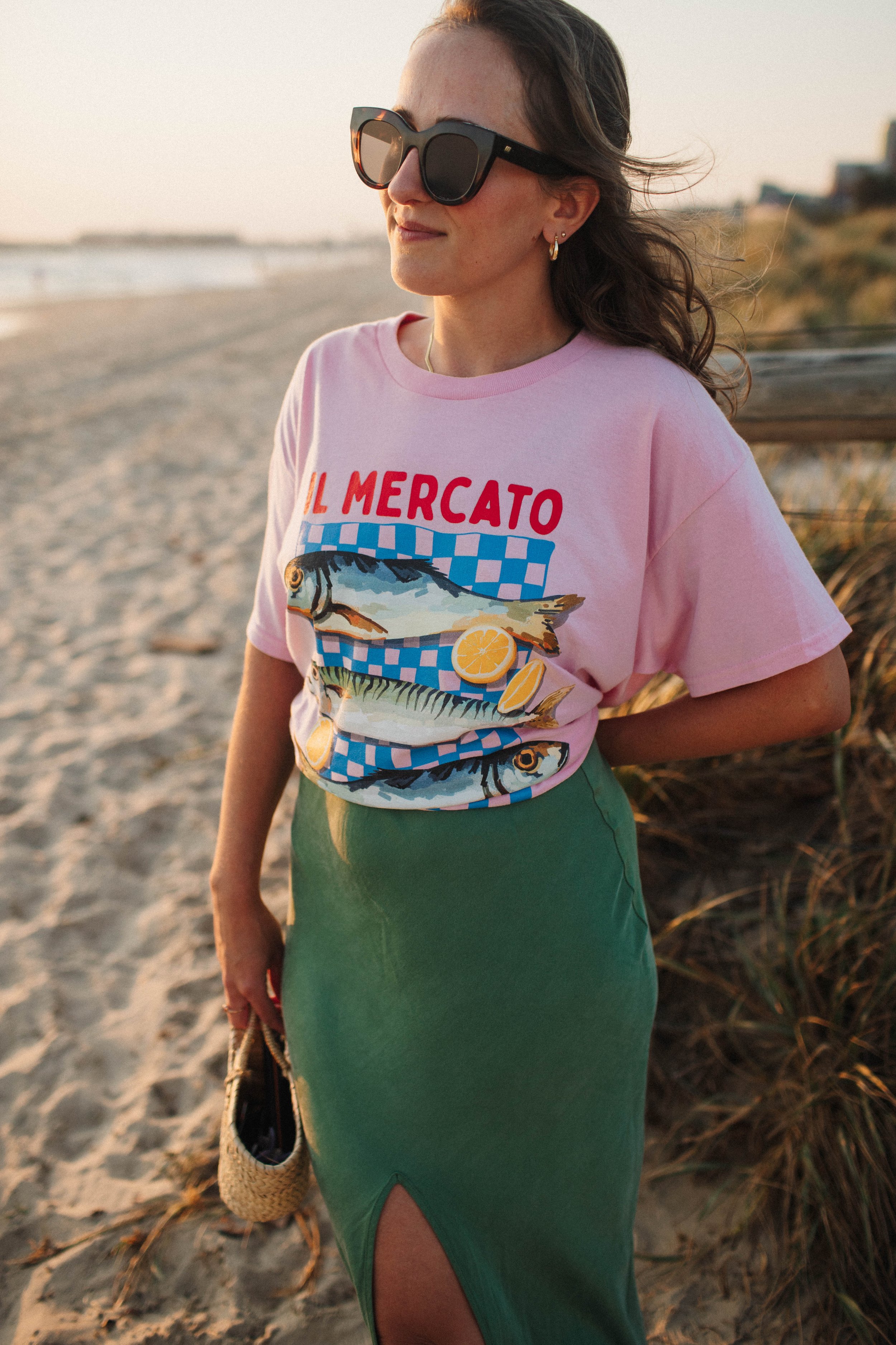 Woman wearing sunglasses and a pink 'Il Mercato' t-shirt with fish design, standing on a beach in a green skirt holding a woven bag.