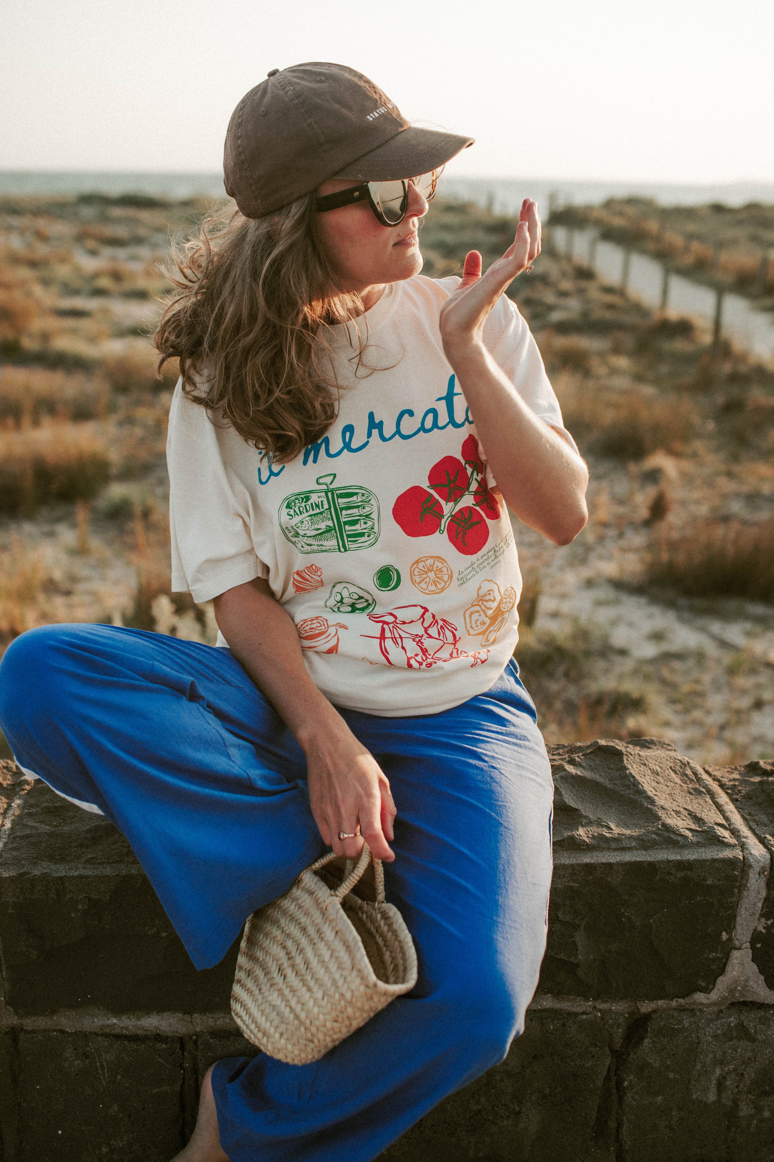 Woman in sunglasses and cap wearing a printed T-shirt and blue pants, sitting on a stone wall with a small wicker bag.