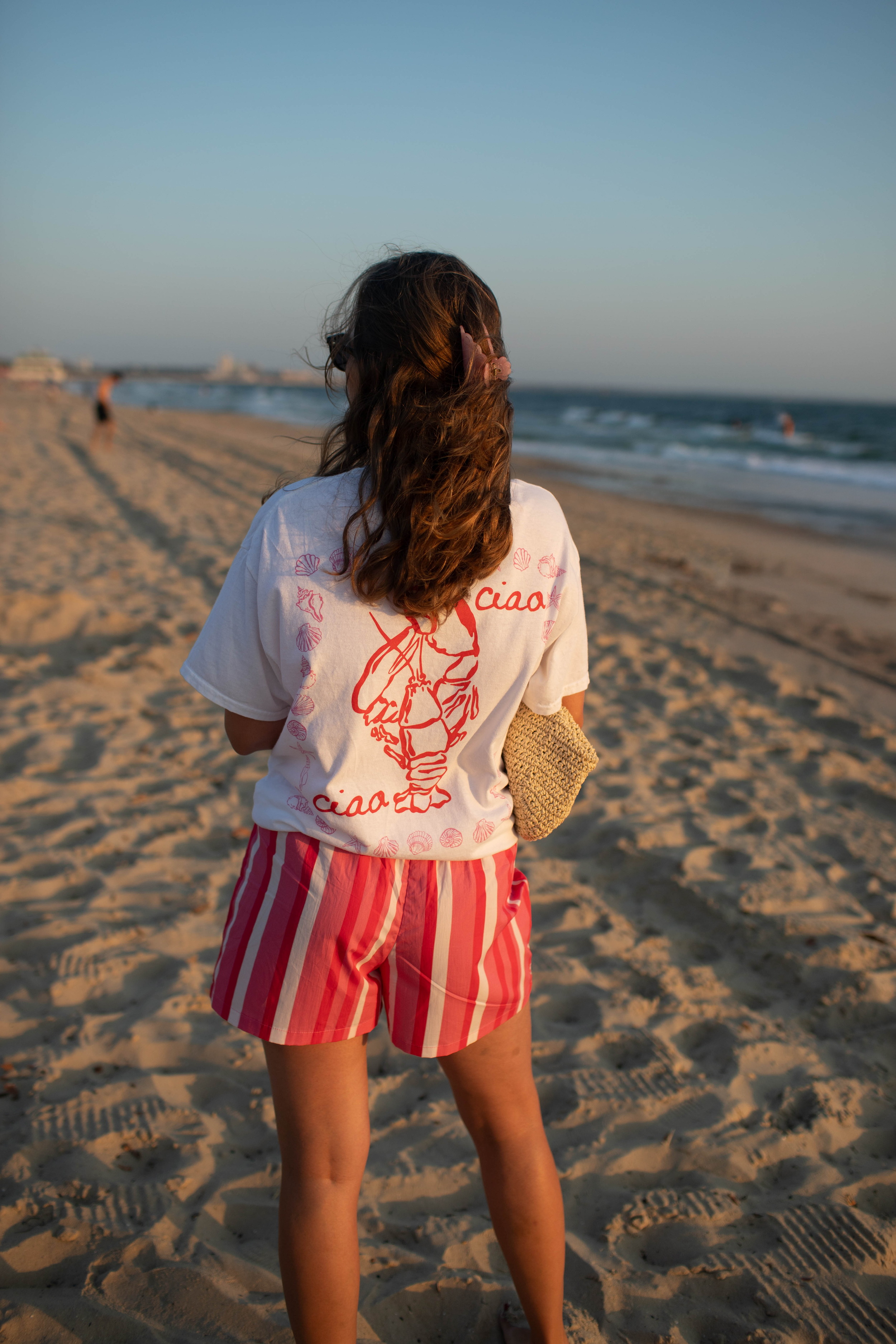 Woman on beach wearing white T-shirt with lobster design and striped shorts, holding a woven bag.