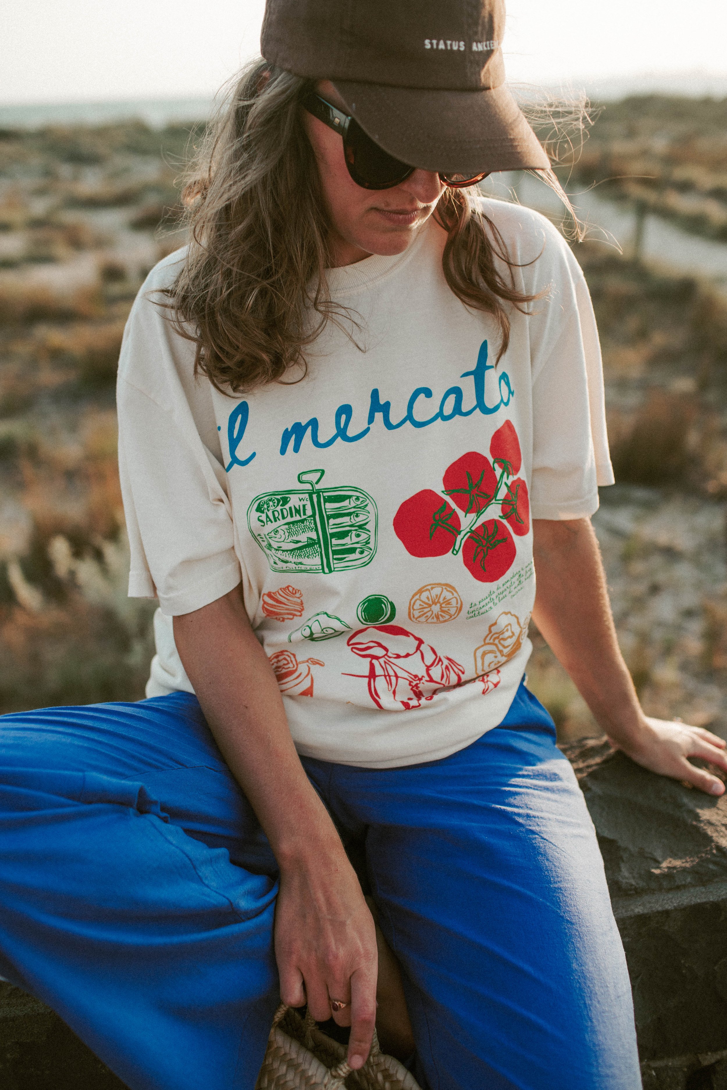 Person wearing a white T-shirt with "il mercato" printed on it, featuring images of sardines, tomatoes, and other food items, sitting outdoors on a stone with a scenic background. Wearing a dark baseball cap and sunglasses, with bright blue pants.