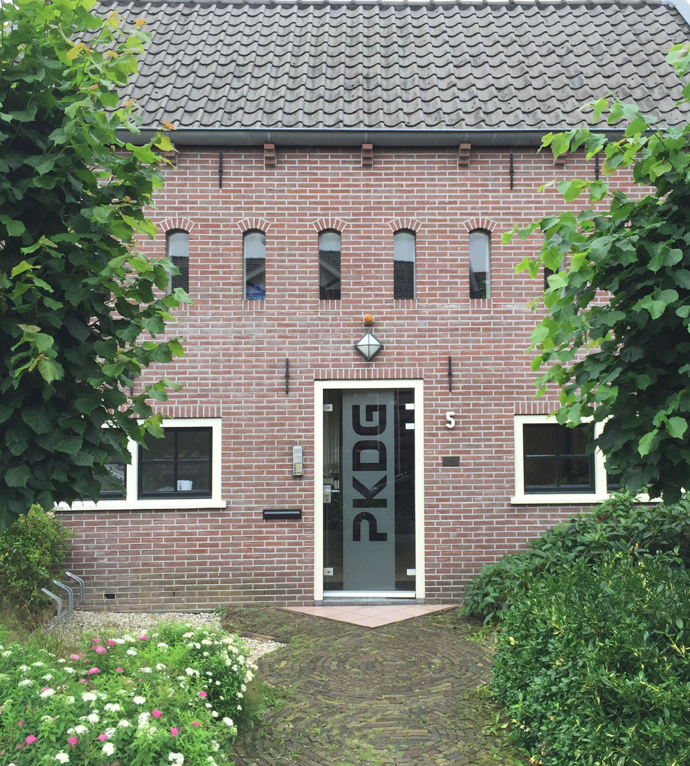 Brick building facade with four vertical windows, arched tops, and a glass door labeled 'PKDG'. The entrance is flanked by small windows and is surrounded by trimmed bushes and flowers.