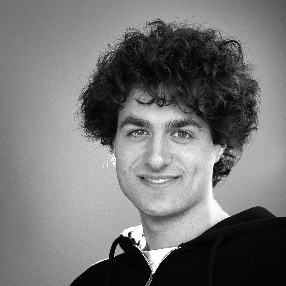 Black and white portrait of a person with curly hair and a hoodie, smiling at the camera.