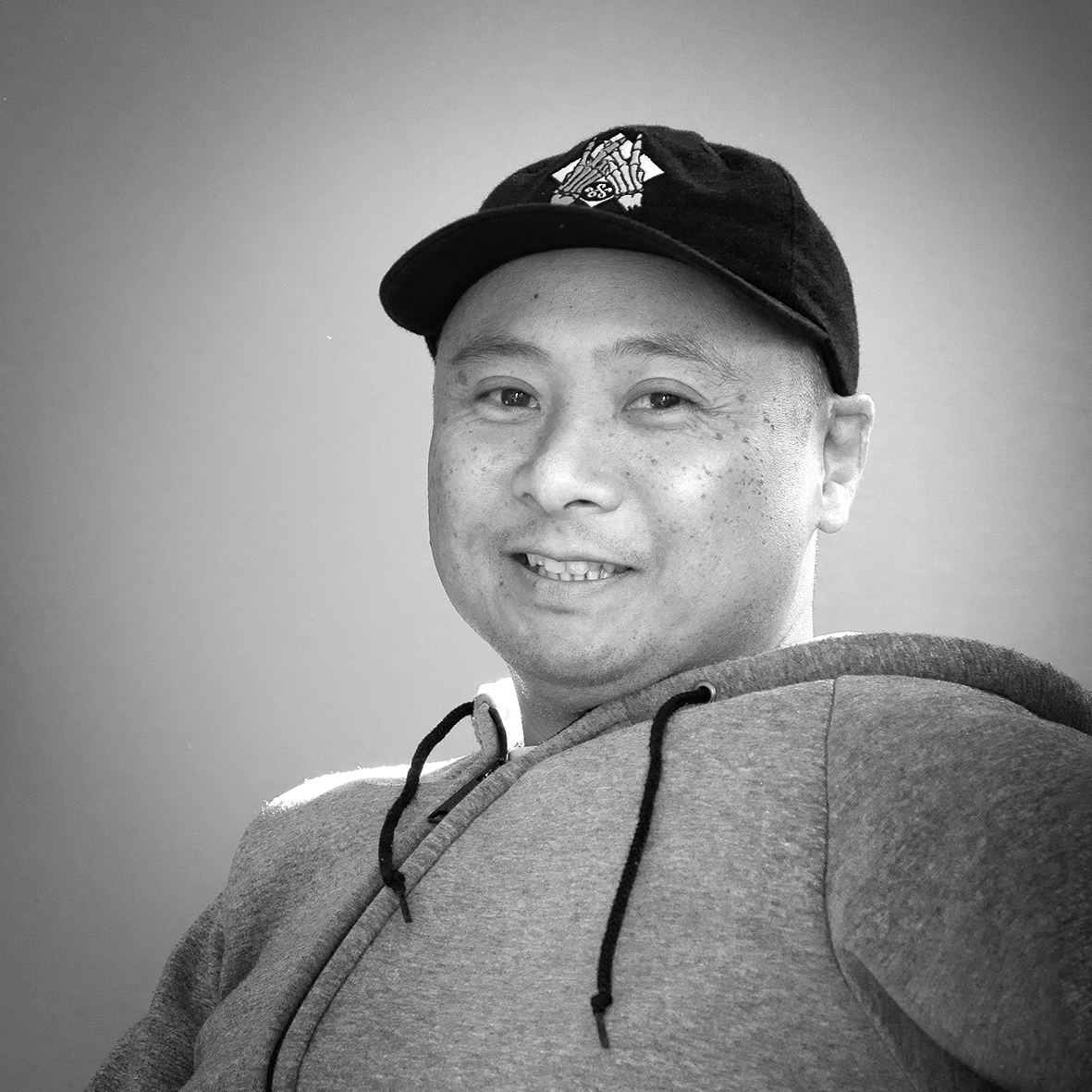 A black and white photo of a person wearing a hoodie and a baseball cap, smiling at the camera.