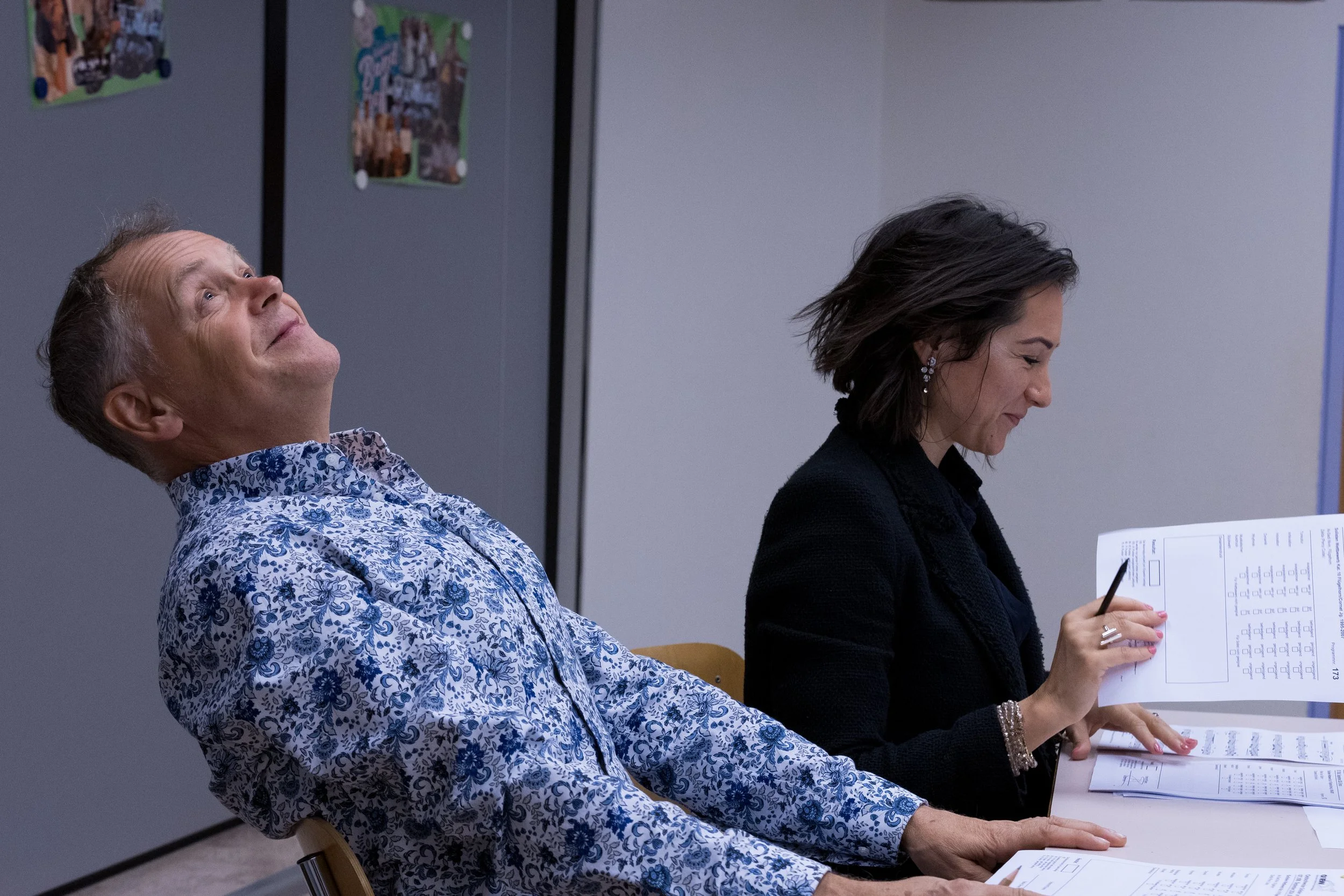 Ein Mann und eine Frau sitzen an einem Tisch, während die Frau ein Dokument durchblättert und den Stift in der Hand hält. Der Mann lehnt mit dem Kopf nach hinten und schaut nach oben.
