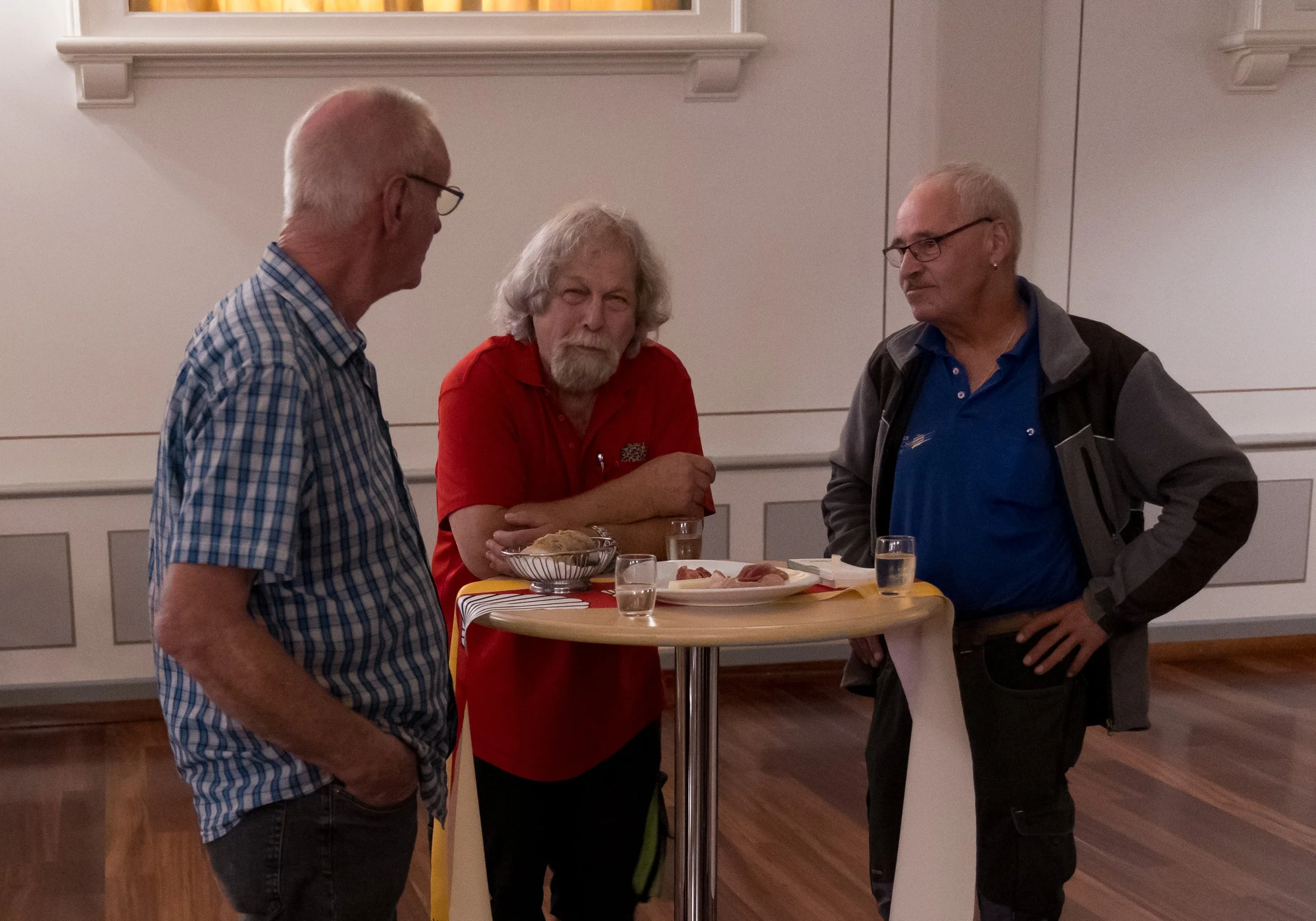 Drei ältere Männer stehen an einem hohen runden Tisch, trinken Schnaps und unterhalten sich in einem Raum mit weißen Wänden und Holzboden.