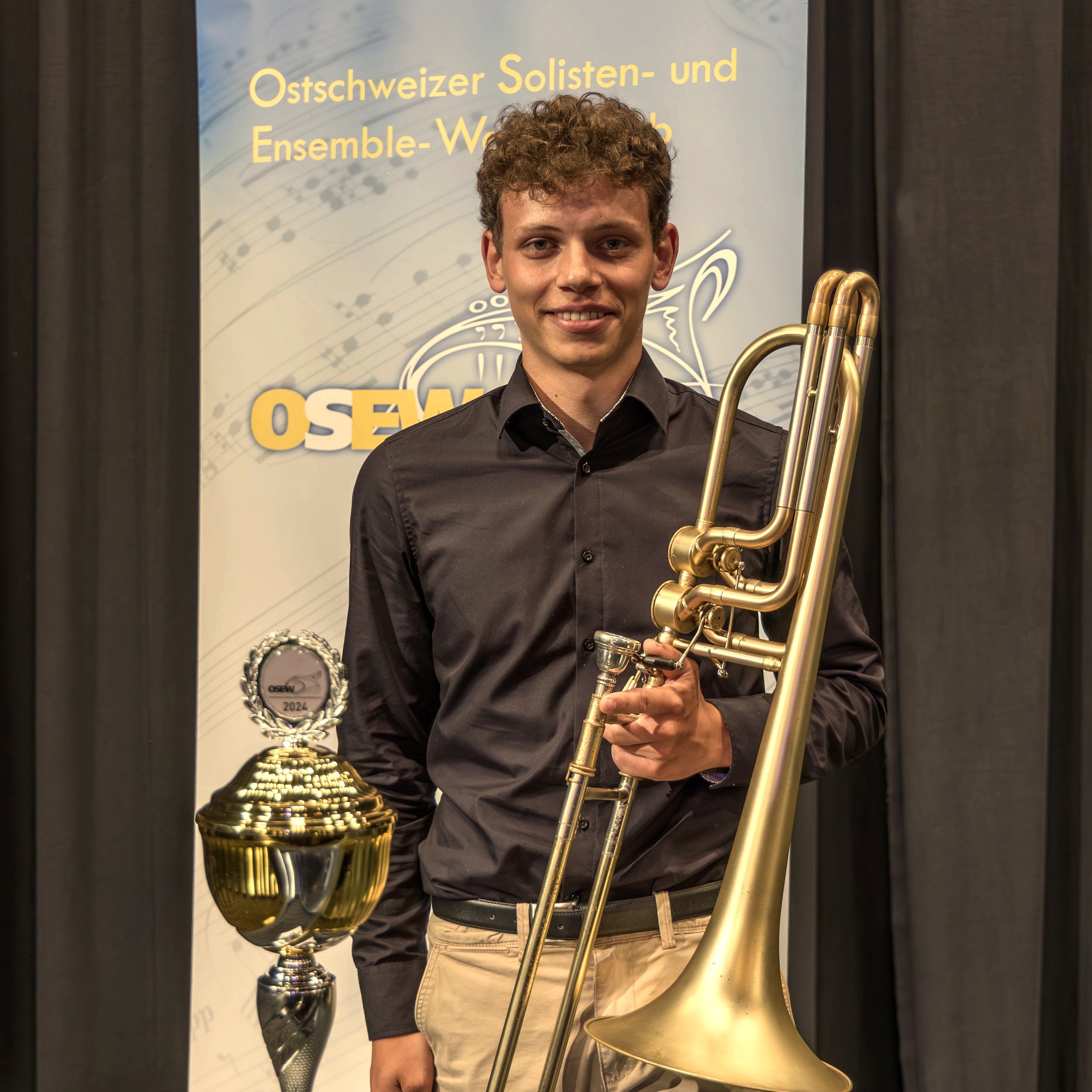 Junger Mann mit hellbraunen, lockigen Haaren, trägt einen schwarzen Hemd, hält eine Bass-Posaune und steht vor einem Banner der OSEW 2024, das einen goldenen Pokal und eine Posaune zeigt.