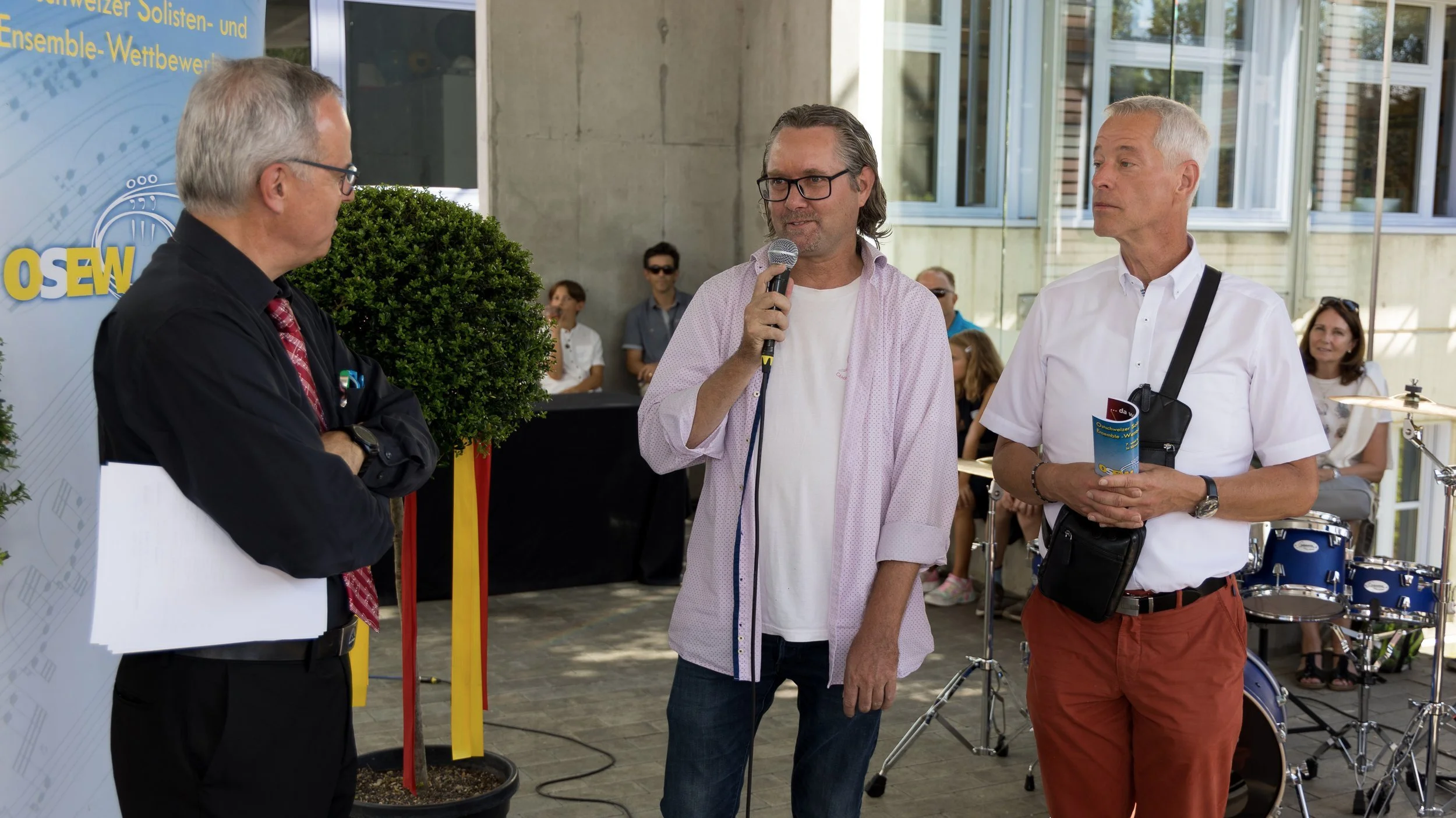 Drei Männer im Gespräch bei einer Veranstaltung, eine Person hält ein Mikrofon, im Hintergrund sind weitere Menschen und eine Drumset. Die Schlagzeugübergabe für Teilnehmende mit grösstem Potenzial.