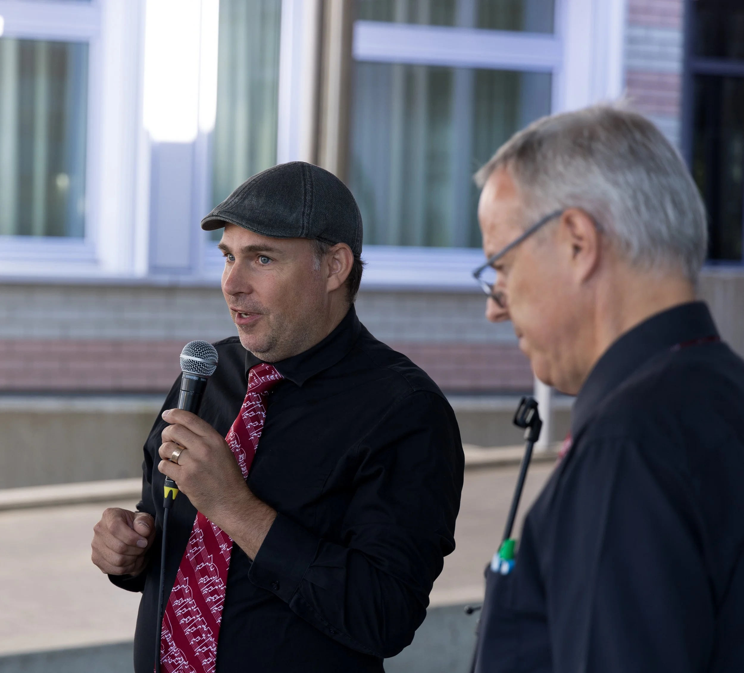 Zwei Männer sprechen bei einer Veranstaltung. Der Mann auf der linken Seite hält ein Mikrofon und trägt eine schwarze Jacke, eine Krawatte mit Musiknoten und einen grauen Kappen. Der Mann auf der rechten Seite trägt eine schwarze Jacke und eine Brille.