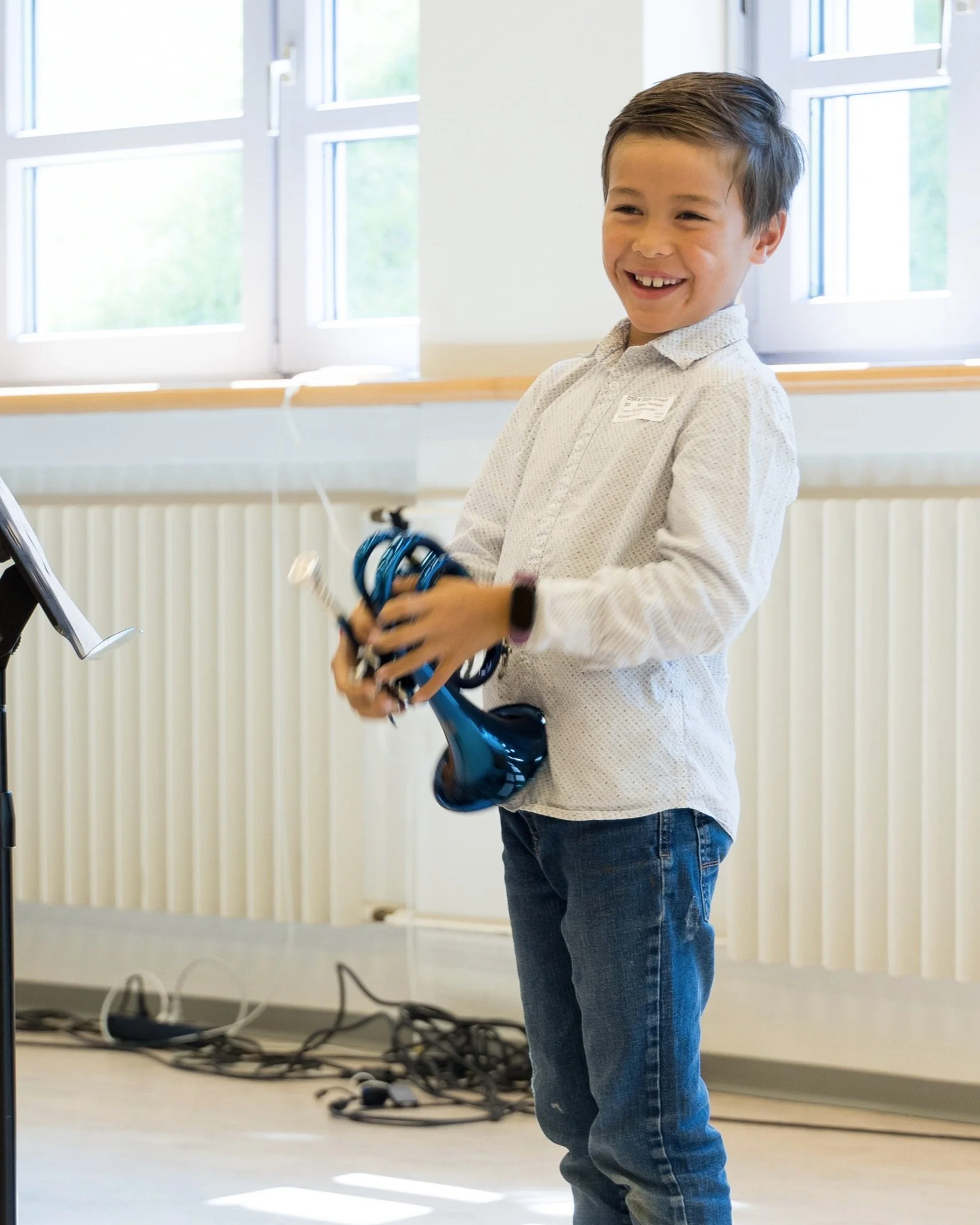 Ein lachender Junge hält ein Kabel und ein Musikinstrument, wahrscheinlich in einem Klassenraum, bei einer zeitgenössischen Musik- oder Schulaktivität.