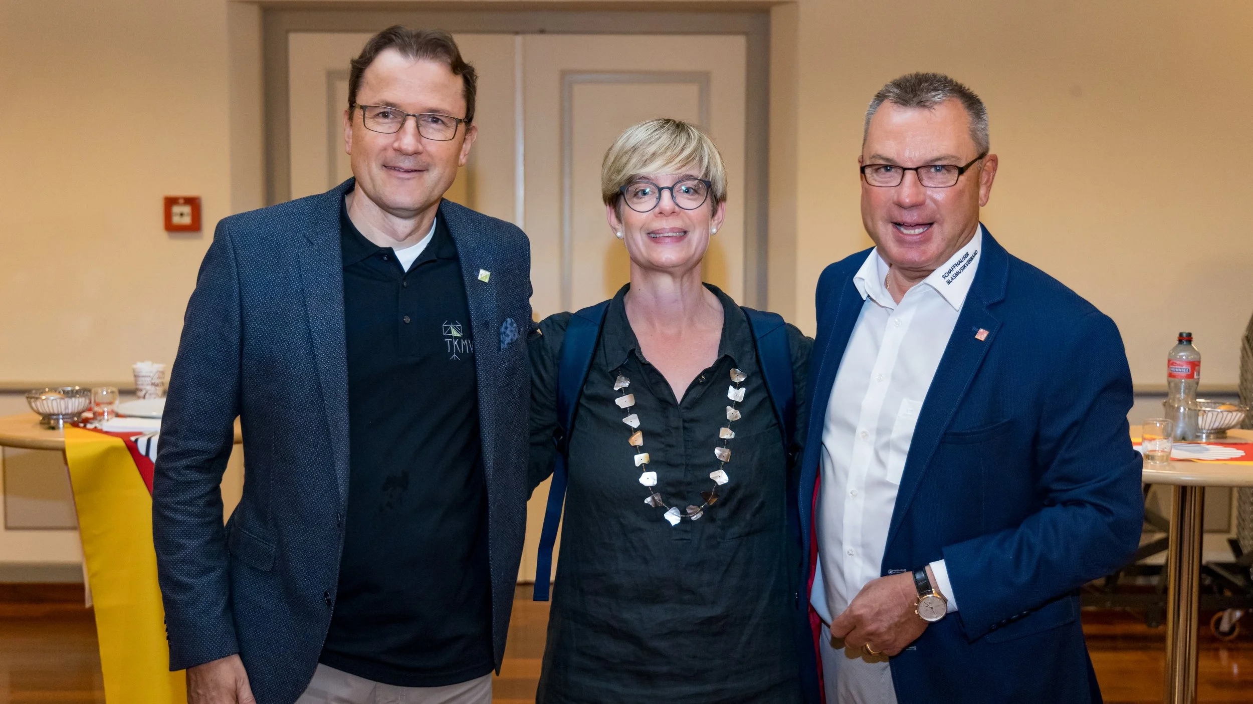 Drei Menschen, zwei Männer und eine Frau, posieren zusammen in einem Raum. Sie tragen formelle Kleidung und lachen. Es gibt Tische im Hintergrund mit Tellern und Getränken.