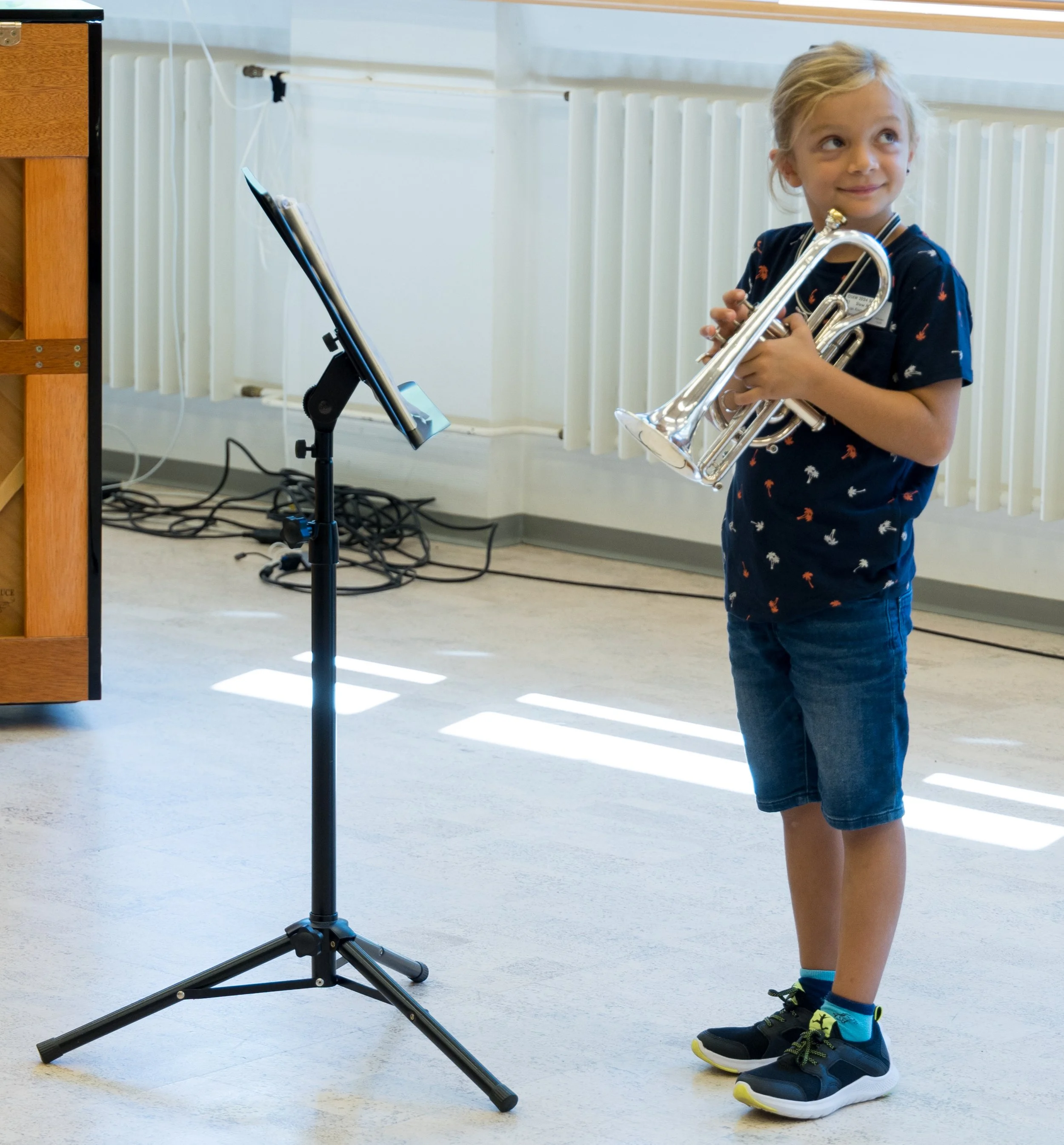Ein Mädchen hält eine Trompete in den Armen, steht vor einem Stativ mit einem Notenständer, im Hintergrund eine Heizung und Kabel auf dem Boden.