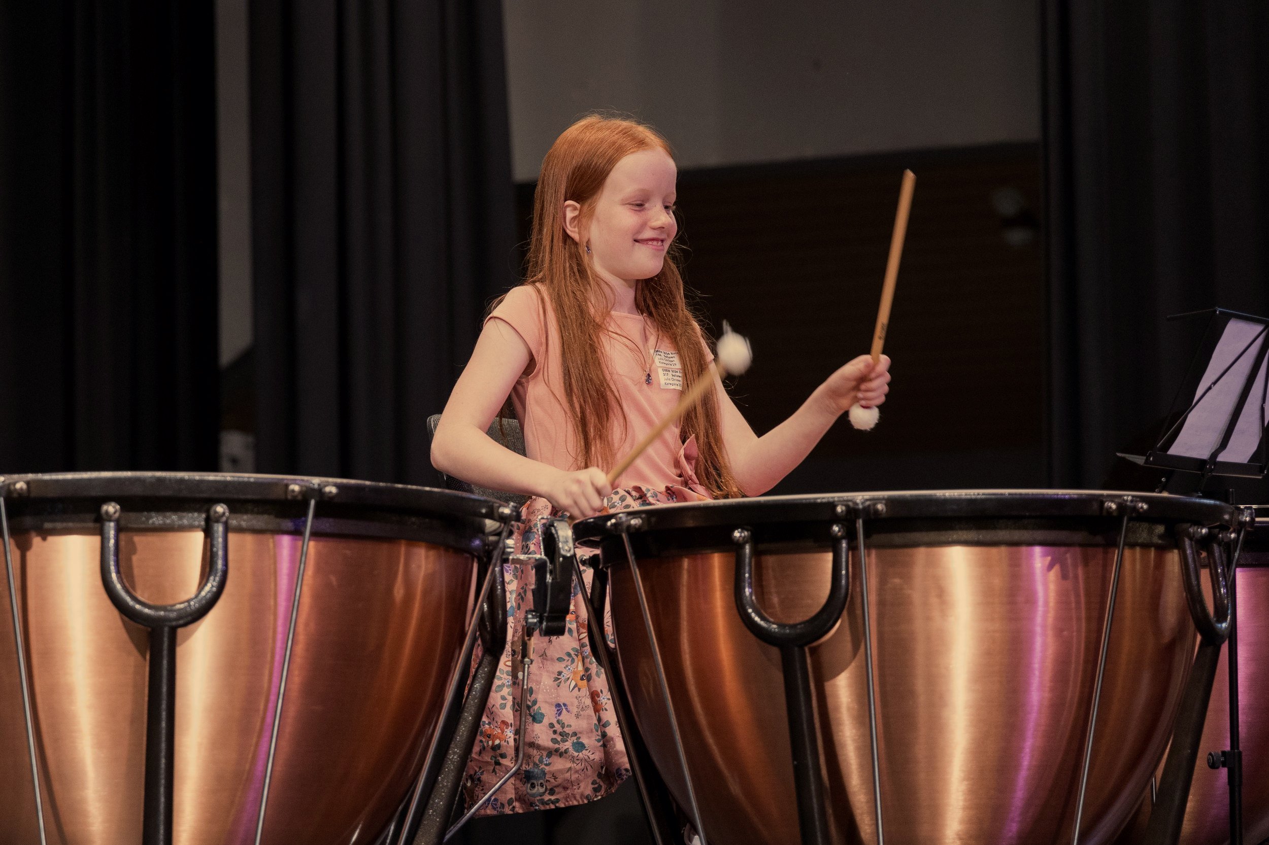 Ein junges Mädchen spielt auf einem großen Perkussionsinstrument, wahrscheinlich eine Marimba oder ein Vibraphon, auf einer Bühne mit schwarzem Vorhang im Hintergrund.